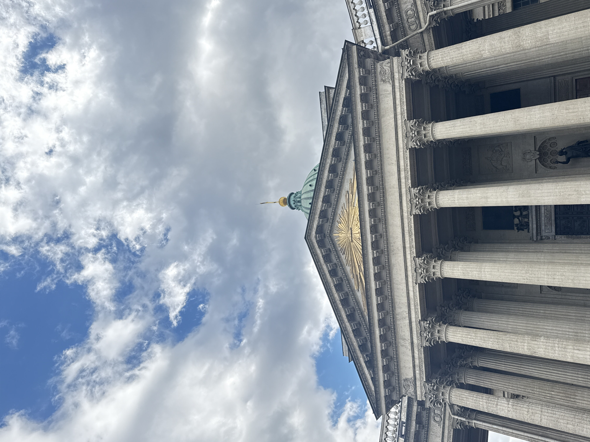 The image shows a low-angle view of the Kazan Cathedral in Saint Petersburg, Russia. The cathedral's facade features a prominent pediment with a gilded radiant sun design, supported by a row of Corinthian columns. The green dome with a golden spire is partially visible above the pediment. The sky in the background is filled with white and gray clouds and patches of blue sky. The building is light grey in color and the architectural style is neoclassical. No people are visible in the frame.