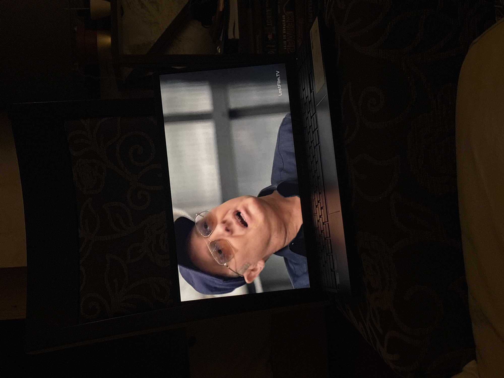 The image displays a screen, likely a laptop or tablet, showing a man's face. The screen is resting on a dark surface with a subtle patterned texture, possibly a table or fabric. To the right of the screen, a portion of a keyboard or the device's base is visible.
On the screen, a man with glasses, wearing a blue cap or hood and a blue top, is depicted with his head tilted upwards. His mouth is wide open, conveying an expression of surprise, laughter, or speaking. In the upper right corner of the displayed content, the text "LostFilm.TV" is clearly visible, identifying the source of the media.
The surrounding environment is dimly lit, suggesting an indoor setting during evening or night hours. Behind the screen, stacked objects resembling books or media cases can be seen. To the far right, a light-colored object, possibly a cushion or bedding, is partially visible. The scene captures an individual viewing content on a personal device in a private, relaxed setting. No specific location cues for Budva, Montenegro are present within the visual content.