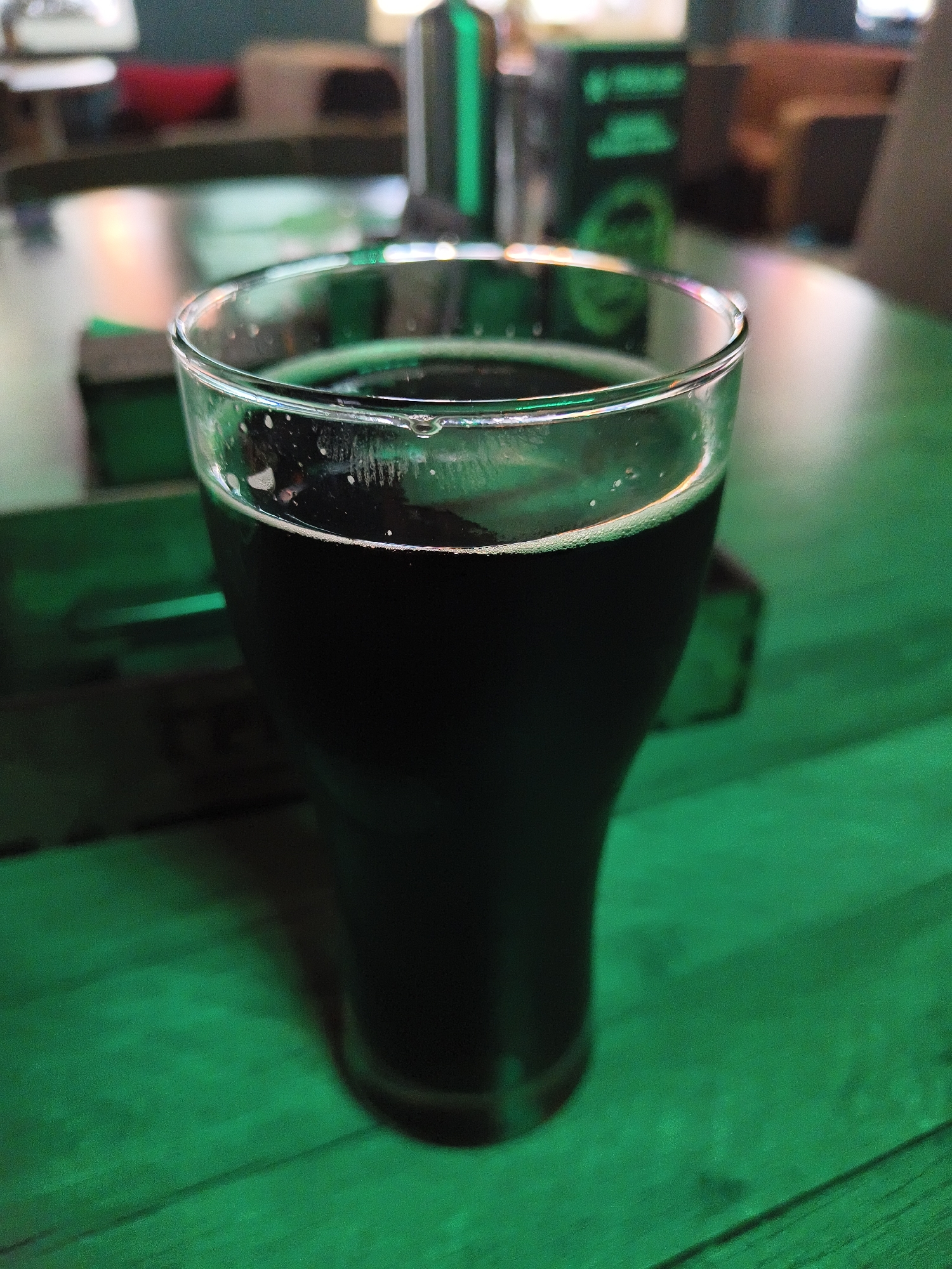 A close-up, high-angle shot focuses on a glass of dark liquid, likely a beverage, with a foamy head, resting on a green-hued wooden surface. The glass is partially filled, and condensation or small droplets are visible on its exterior rim. The background is blurred, showing hints of a dimly lit, indoor environment with green lighting. Several dark rectangular objects are visible in the background, possibly coasters or placemats, also illuminated by the green light. The overall ambiance suggests a bar or a dimly lit recreational space.