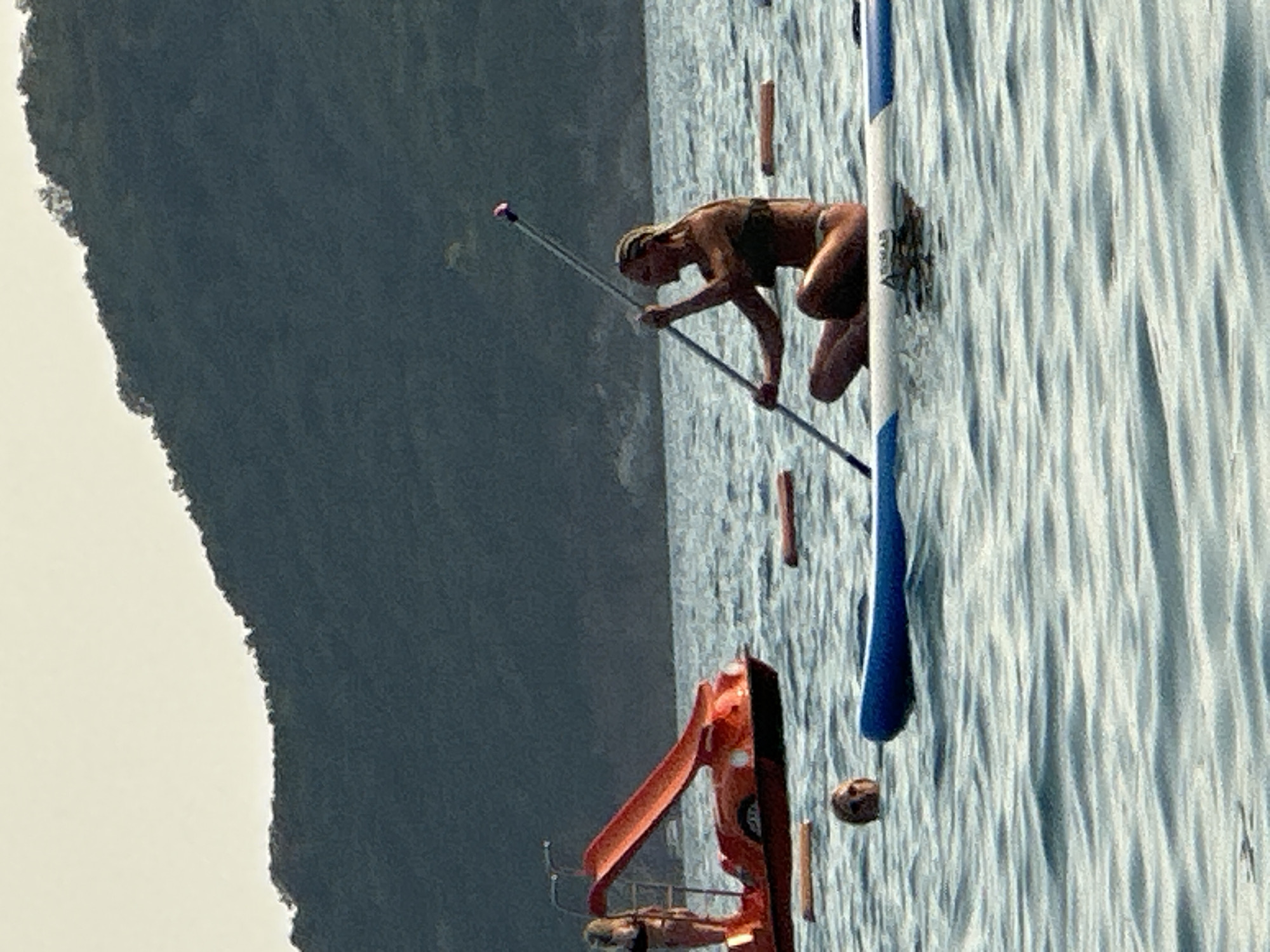 An outdoor, medium shot captures a scenic view of the Boka Bay in Boreti, Montenegro, with a focus on water activities. In the foreground, a woman with blonde hair pulled back is sitting cross-legged on a paddleboard, holding a paddle with both hands. She is wearing a dark bikini. The paddleboard has blue trim. To the left, a portion of another paddleboard is visible. Further in the background, a bright orange pedal boat with a slide is in the water. A person with blonde hair is standing on the pedal boat. The sea is calm with ripples. In the far background, a dark, tree-covered hill slopes down towards the water under a hazy sky. Two brown, cylindrical buoys are floating in the water.