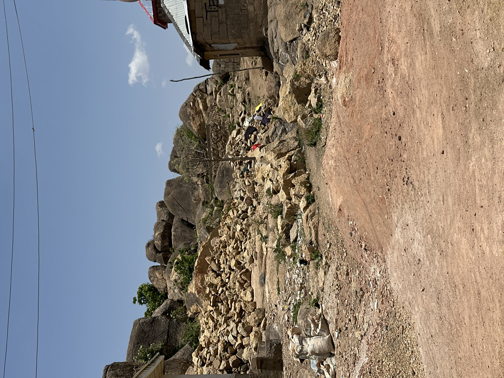 The image depicts a rocky hillside with a dirt path alongside, likely in Jos, Nigeria, given the terrain. The hillside is covered in boulders and smaller rocks, with sparse vegetation. A small, simple building sits atop the rocks. Clothes are strewn on the rocks. The sky is blue with some clouds. A power line is visible. Debris litters the base of the hillside along the dirt path.
