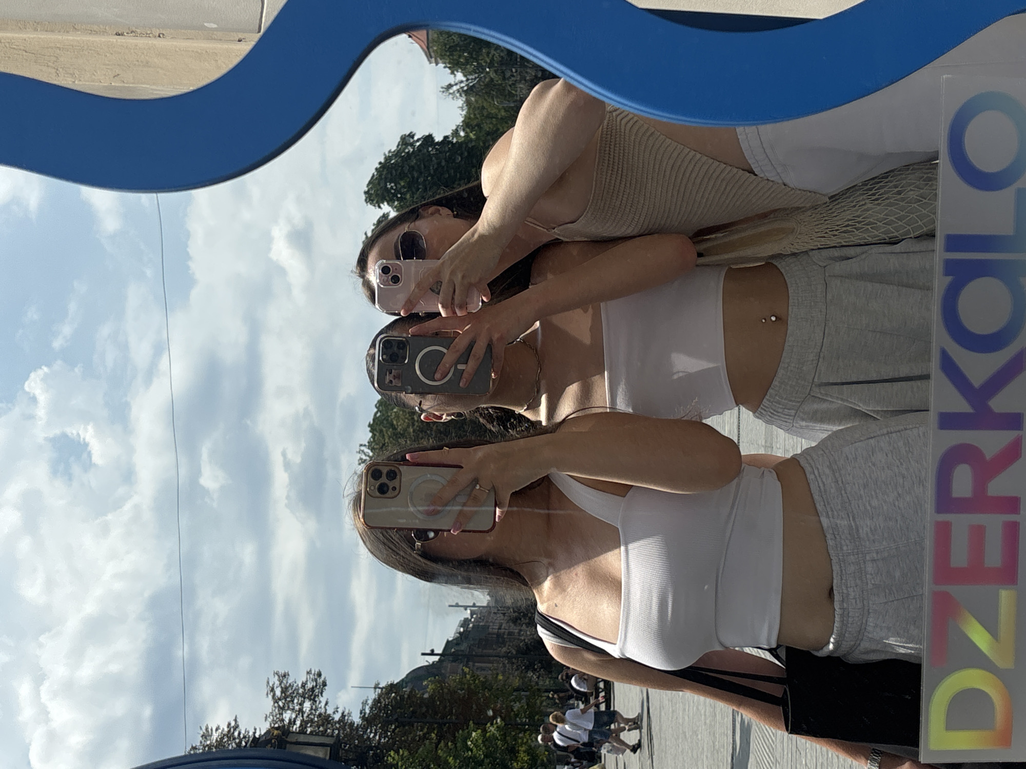 The image depicts a mirror reflection of three young women standing outdoors, possibly in a city square. The woman on the left is wearing a white tank top, grey sweatpants, and sunglasses. She is holding a smartphone to take a selfie. To her right, another woman in a white crop top and grey sweatpants is also holding a smartphone. The woman on the far right is wearing a beige knit top and white pants, also holding a smartphone. In the background, through the mirror, a street scene is visible with people walking, trees, and buildings. A blue, wave-like frame surrounds the right side of the mirror. At the bottom of the image, the colorful word "DZERKALO" is visible, which is Ukrainian for "mirror," suggesting the photo was taken in Lviv, Ukraine. The sky is cloudy.