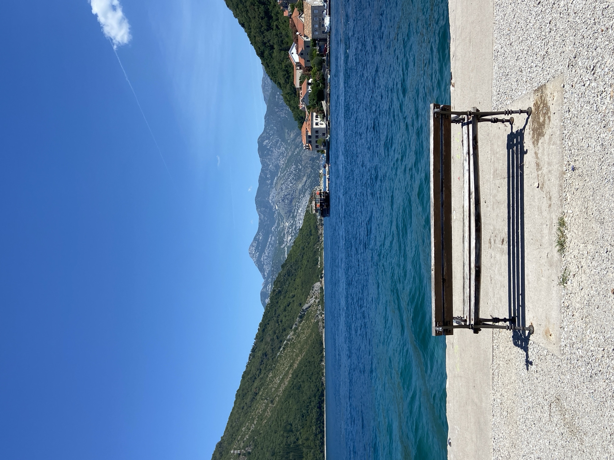 In Lepetane, Montenegro, on a clear, sunny day, a lone wooden bench with a dark metal frame sits on a concrete and gravel walkway. The bench faces a tranquil, turquoise body of water, likely the Bay of Kotor, with gentle waves lapping the shore. In the distance, lush green mountains rise from the water's edge, their slopes dotted with trees and rocky outcrops. A small cluster of buildings with terracotta roofs is visible on the far side of the bay, nestled at the foot of the mountains. A ferry boat is docked near the buildings, and other smaller vessels are scattered across the water. The sky is a vibrant blue with a few wispy clouds and a faint contrail from an aircraft. The scene evokes a sense of peaceful serenity and natural beauty.