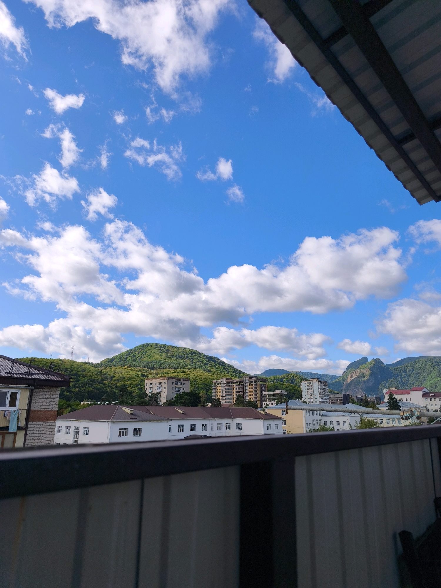 The image shows a view of buildings and mountains under a blue sky with scattered white clouds. In the foreground, a dark metal railing obscures the bottom portion of the scene. Behind the railing, a white building with a brown roof is visible, with several windows. To the left, a brick building with a brown roof is partially visible, with laundry hanging from a window. Further in the background, a row of multi-story buildings can be seen against a backdrop of green, tree-covered mountains. In the far distance, rocky mountain peaks are visible. The sky is bright blue with fluffy white clouds. The scene appears to be a residential area within a mountainous region.