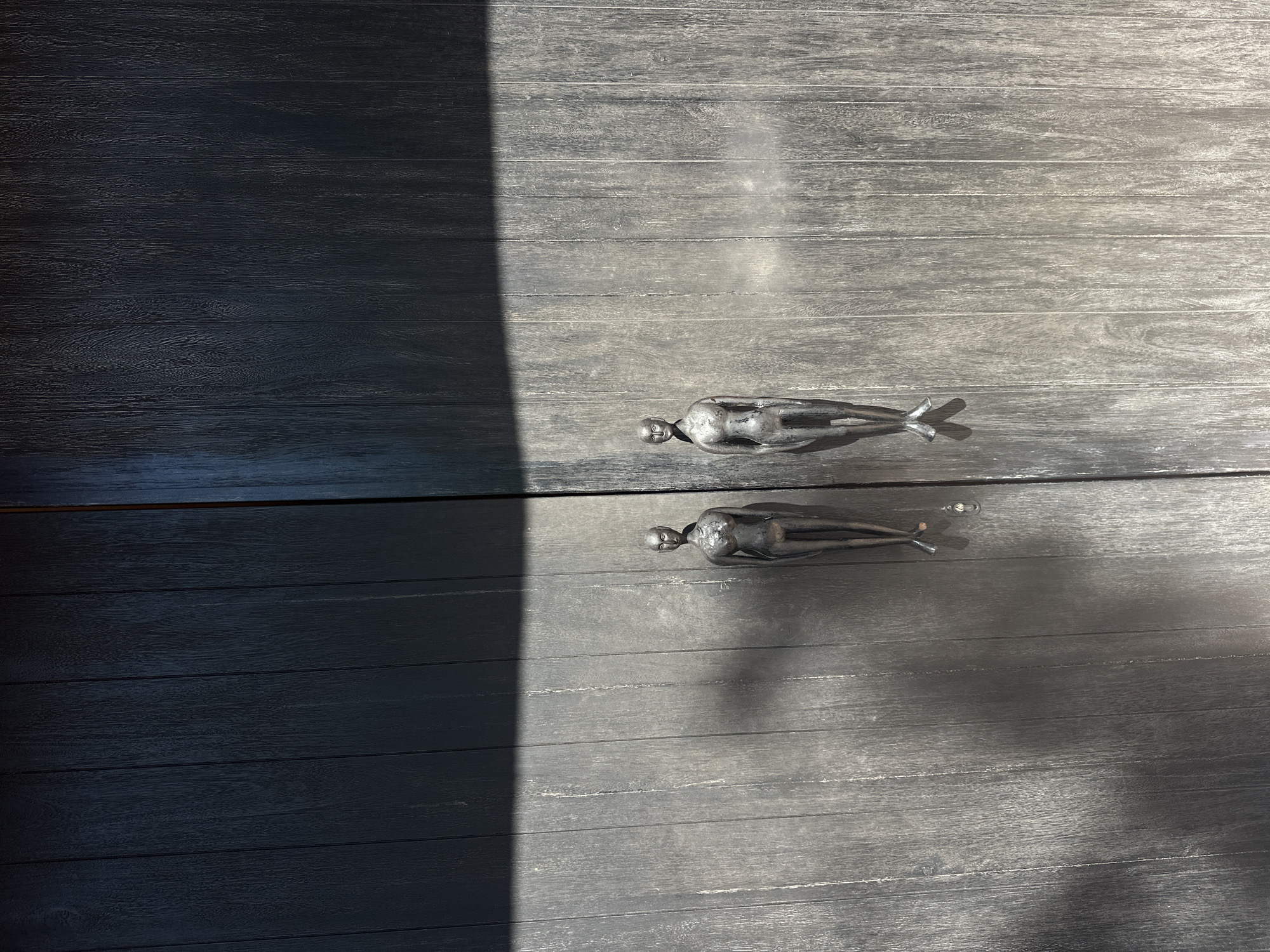 The image shows a weathered grey wooden surface with two decorative metal figurines. The figures appear to be stylized human forms, oriented horizontally. One figurine is on the upper half of the image, and the other is directly below it on the lower half of the image. A stark diagonal line separates a sunlit portion of the wooden surface from a darker, shadowed area. The scene's location is speculated to be in Pecatu, Indonesia, based on contextual information.