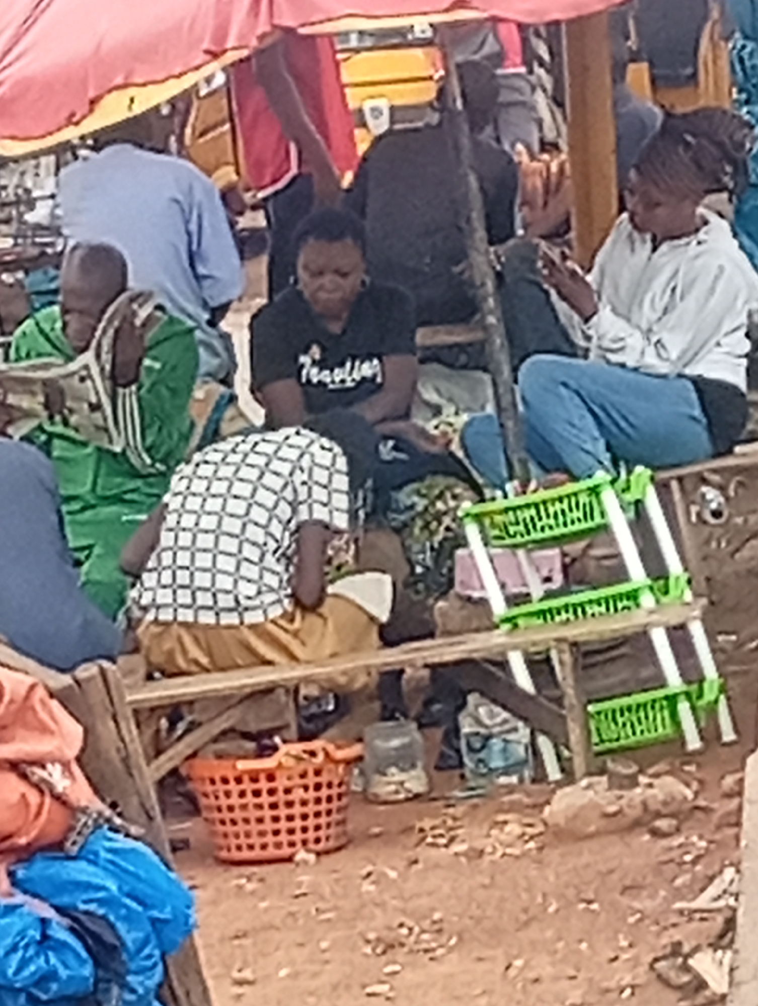 The image depicts a market scene in Sabon Tasha, Nigeria. Several individuals are visible, some sitting on wooden benches. One woman wears a black T-shirt and sits with her arms crossed. Another woman, wearing a white hoodie and blue jeans, sits with legs crossed, looking at a mobile phone. A man in green clothing sits to the left. A person in a checkered shirt and mustard-colored pants sits facing away from the camera. Underneath the seating area is an orange plastic basket and a green plastic shelving unit. A canopy is visible overhead. The ground is brown and appears to be dirt.