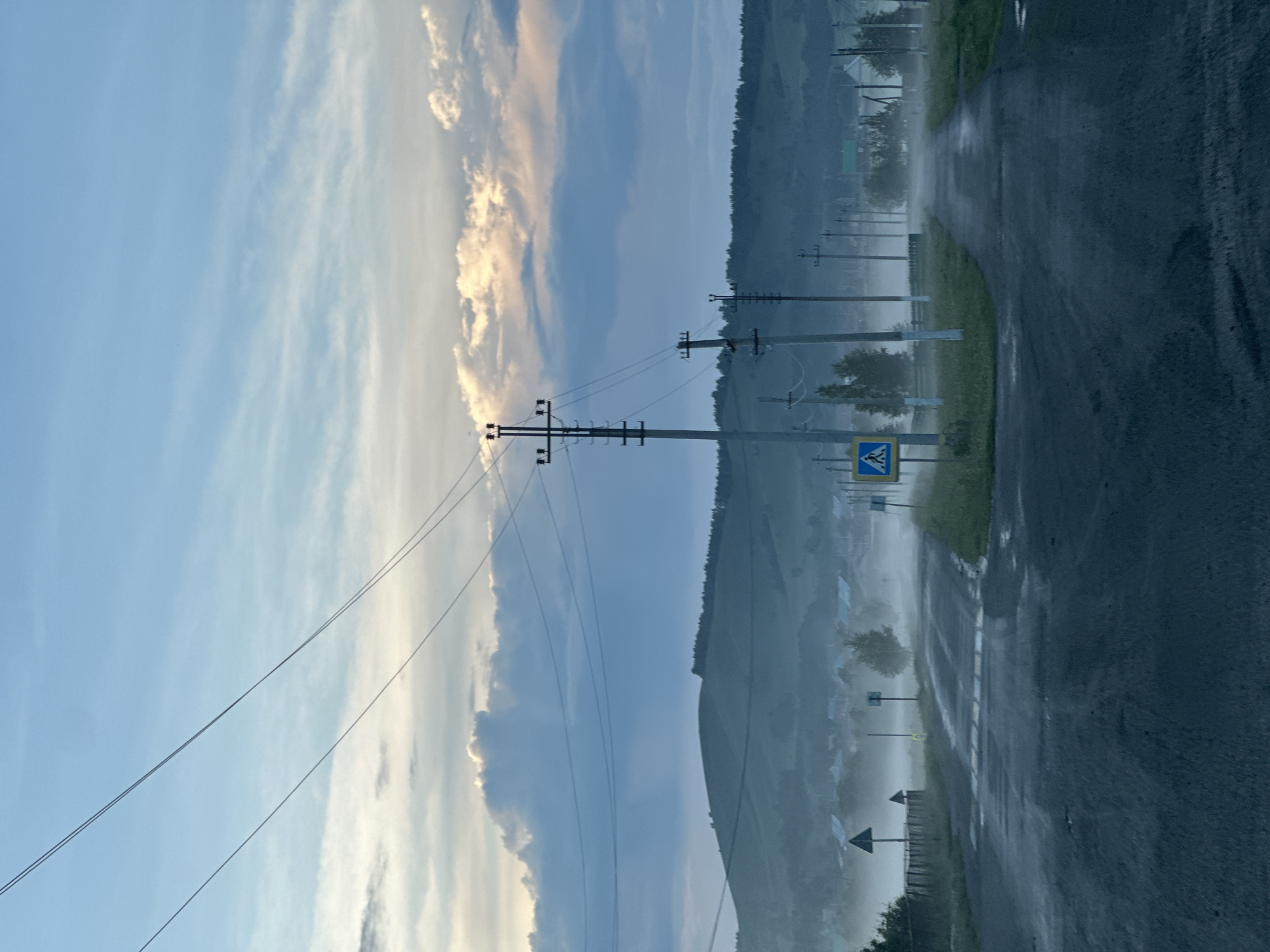 This image depicts a rural or semi-rural road intersection at dawn or dusk in the city of Unknown, Unknown. The scene is characterized by dense ground fog or mist, a wet asphalt road, and overhead power lines.
The sky in the upper portion of the image is light blue with white and gray stratiform clouds. Towards the horizon, the clouds exhibit faint golden highlights, indicating either sunrise or sunset. Several thin, dark power lines traverse the upper sky diagonally.
The midground features undulating hills covered in vegetation, some densely forested with coniferous trees, particularly on the right side. At the base of these hills, a small settlement or scattered houses are visible, partially obscured by the heavy mist. Numerous utility poles, carrying multiple wires, extend vertically into the frame and recede along the road into the distance.
In the foreground, a dark, wet asphalt road forms a junction. The road surface shows signs of moisture, including puddles and reflective sheen. A pedestrian crossing, marked by white stripes, is visible on the horizontal road segment. A blue and yellow pedestrian crossing sign stands on the left side of the road at the junction. Another similar sign is partially visible on the right. Green grass verges line the sides of the road. No individuals, animals, or vehicles are present in the frame. The overall atmosphere is cool and muted, dominated by blues, grays, and greens, with diffused light filtering through the misty conditions.