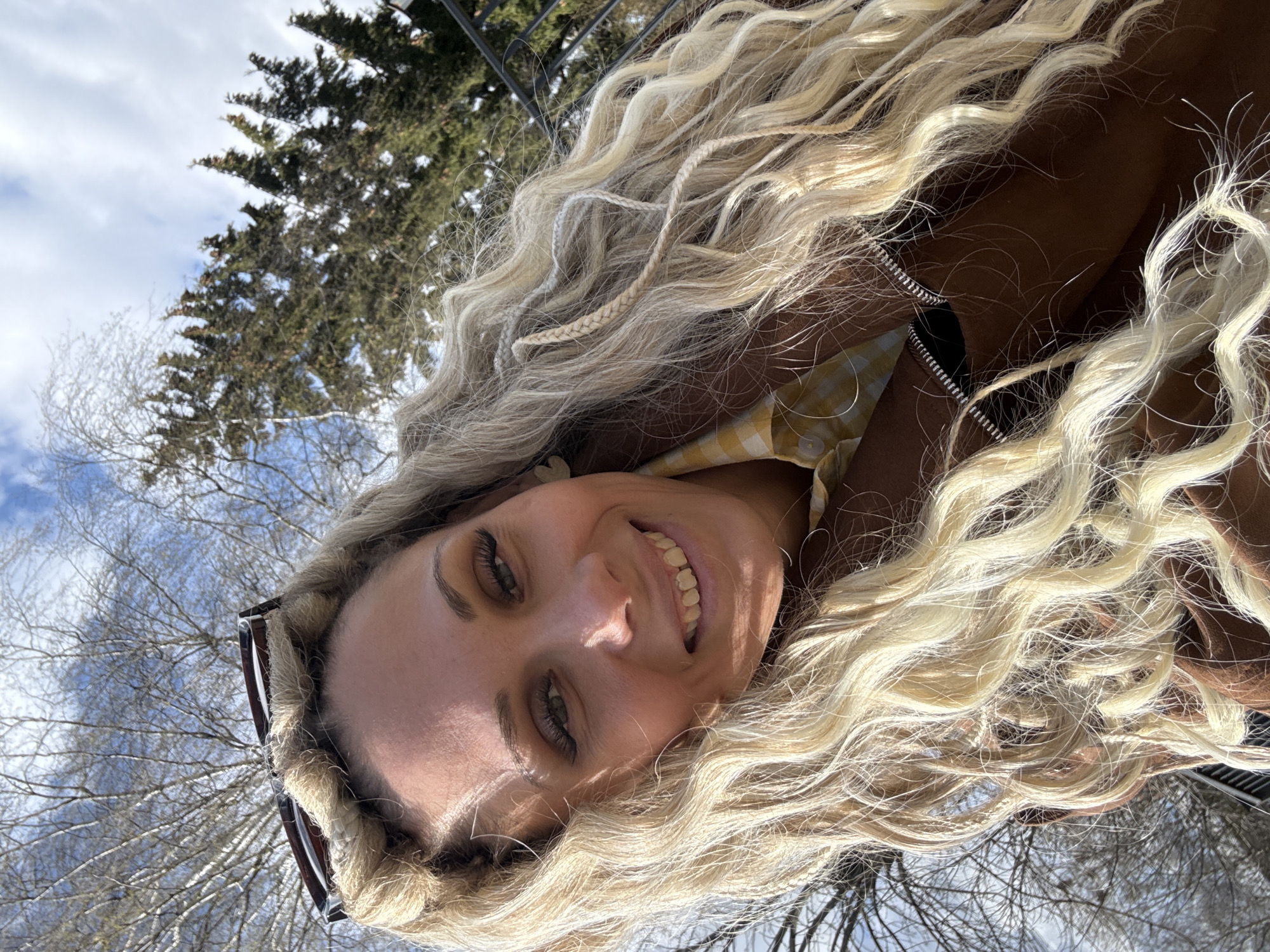 A woman with long blonde curly hair is posing for a selfie outdoors. She is smiling, and her sunglasses are perched on top of her head. She is wearing a brown jacket over a yellow gingham shirt. In the background are bare trees against a blue sky with clouds, and a coniferous tree is also visible. The location may be in Rusanovo, Russia.