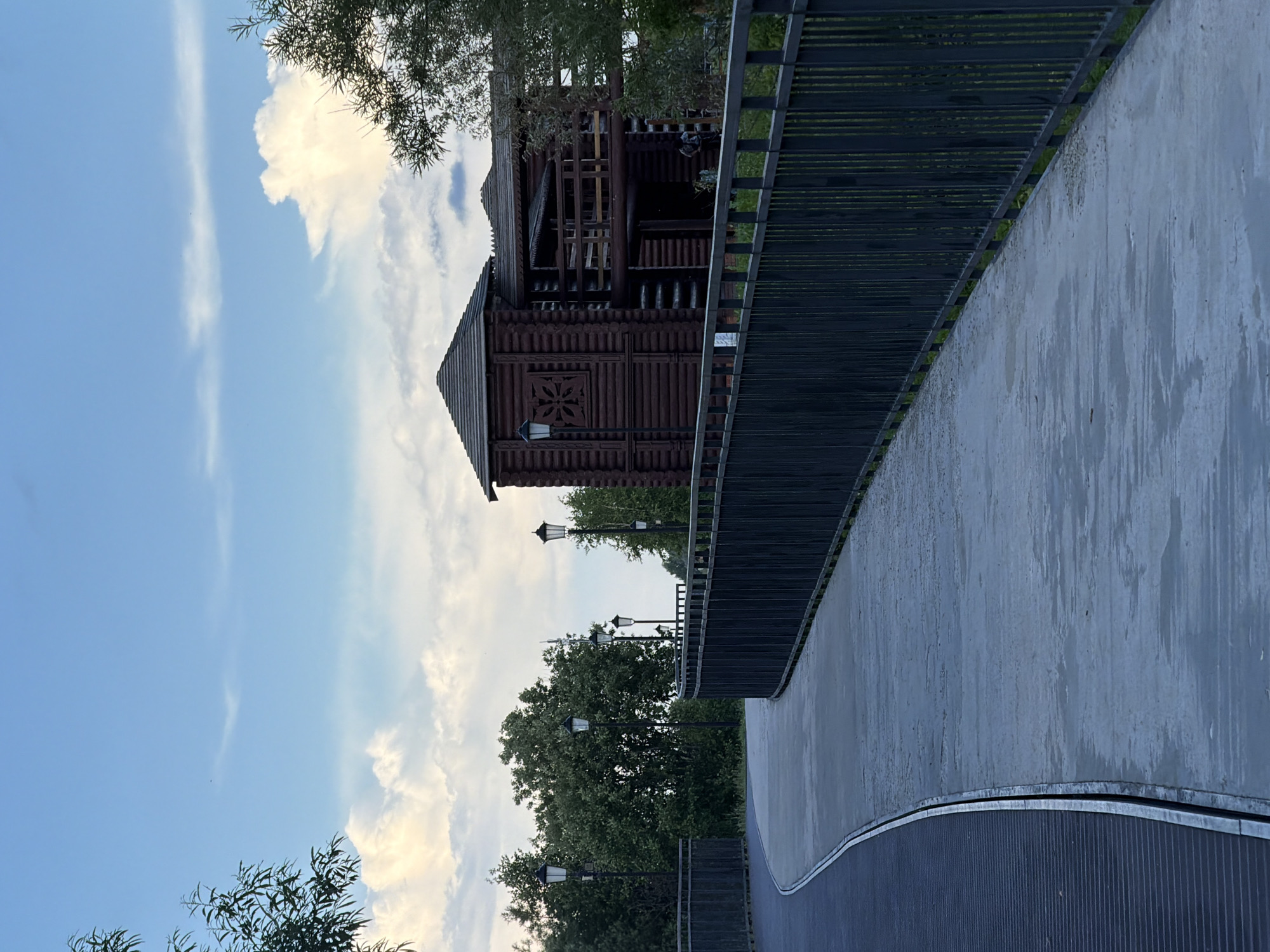 The image depicts a paved pathway curving to the right, bordered by a dark metal fence. On the left side of the pathway, there is a dark metal surface with horizontal slats. Beyond the fence, on the right side of the pathway, a wooden building with a distinctive architectural style, possibly resembling a traditional Russian dwelling or a historical fortification, is visible. This structure is made of dark logs and features multiple roofs, balconies, and decorative elements. The pathway is lined with several black lampposts, some of which are illuminated. In the background, lush green trees form a dense foliage. The sky above is a light blue with scattered white clouds, suggesting it is daytime. The overall setting appears to be an outdoor recreational area or park. This location is identified as being in the city of Solikamsk, Russia. No specific actions or interactions are discernible, and no individuals are prominently featured or identifiable.