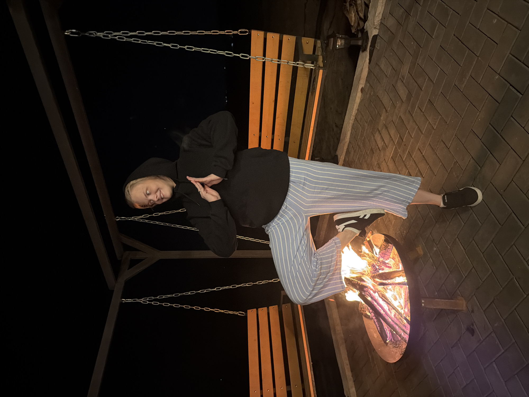 A person is sitting on a hanging bench swing near a fire pit. The person is wearing a black hooded sweatshirt and blue and white striped pants. The fire pit is filled with burning wood. The area around the fire pit is paved with brown bricks. The photograph was taken outdoors at night. The location is Kamennoye, Russia.