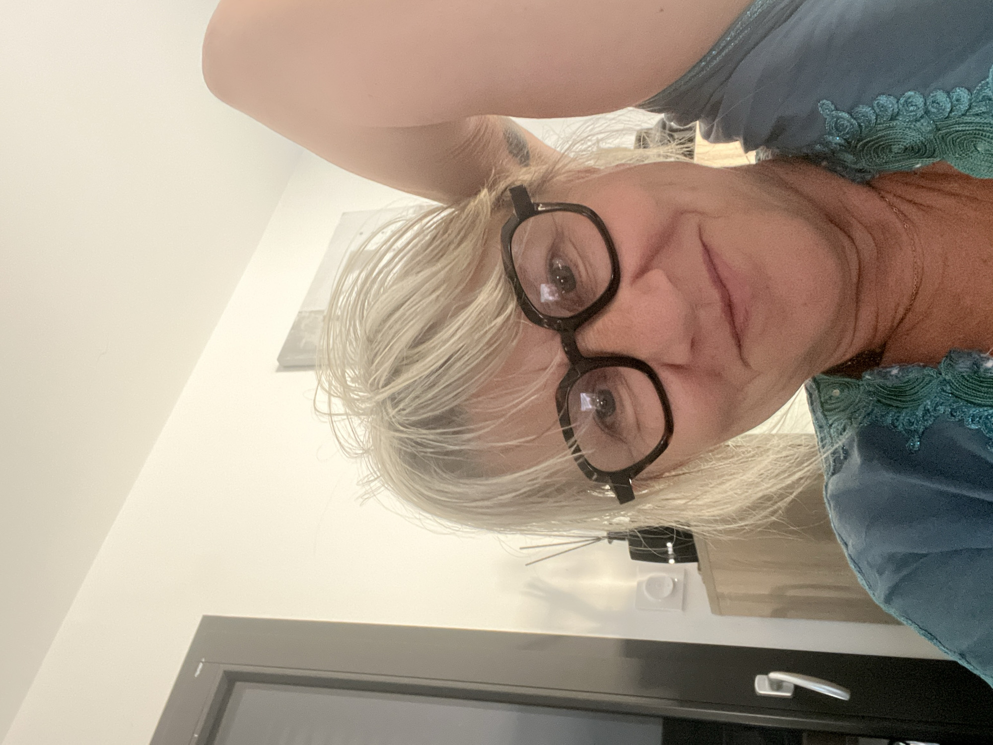 A blonde woman with glasses is taking a selfie in a room. She is wearing a blue top with green trim. Her arm is raised behind her head. A door and a cabinet are visible in the background. The image does not contain enough information to determine the location in Chambœuf, France.