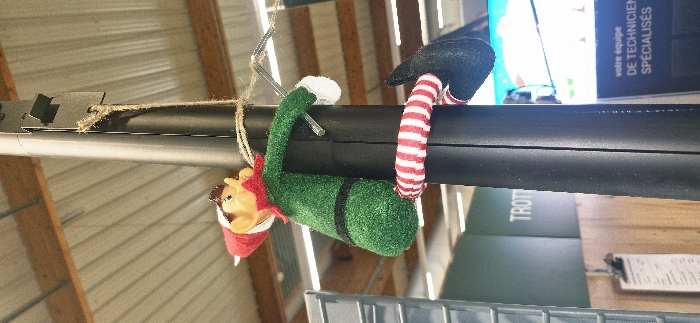 An elf-like plush toy is prominently displayed, hanging from a dark cylindrical structure in an indoor commercial or industrial setting in Vendin-le-Vieil, France. The toy is predominantly green, featuring a red pointed hat, pointed ears, and a light-colored face. Its legs are distinctively striped in red and white.
The activity depicted is the elf toy "climbing" or "hanging" from the cylindrical object. Its arms are wrapped around the cylinder, with one arm appearing to grasp a small, reflective metal hook or piece. Its striped legs are also curled around the cylinder, reinforcing the impression of playful suspension.
Notable details include a piece of brown twine securely tied around the cylindrical object near the elf's head, and a metal bracket situated close to the twine. The background consists of a light-colored, corrugated metal ceiling and visible wooden support beams, suggesting a functional space like a warehouse or workshop. In the deeper background, a blue screen or poster is visible, displaying French text that reads: "votre équipe de TECHNICIENS SPÉCIALISÉS" (your team of SPECIALIZED TECHNICIANS). Below this, a partially visible green object has the word "TROT" printed on it, and another lighter object, possibly a clipboard, is also present. The overall scene, captured in Vendin-le-Vieil, France, integrates a whimsical decoration within an otherwise practical environment.