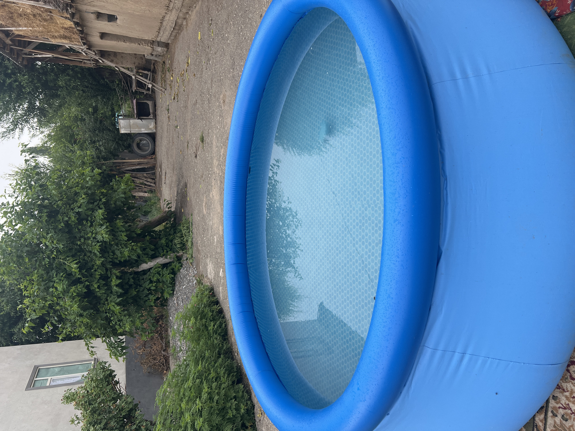 An inflatable blue pool filled with water sits in the foreground of an outdoor yard. The pool has a textured blue liner at the bottom with a hexagonal pattern. In the background, there is a light-colored building with a window on the left, and a rustic structure with a thatched roof on the right. A tire and what appears to be a discarded appliance are visible in the yard between the buildings. Green trees and bushes surround the area. The ground is covered with gravel and dirt. The scene is likely located in Kara-Dıykan, Kyrgyzstan, given the architectural style and vegetation.