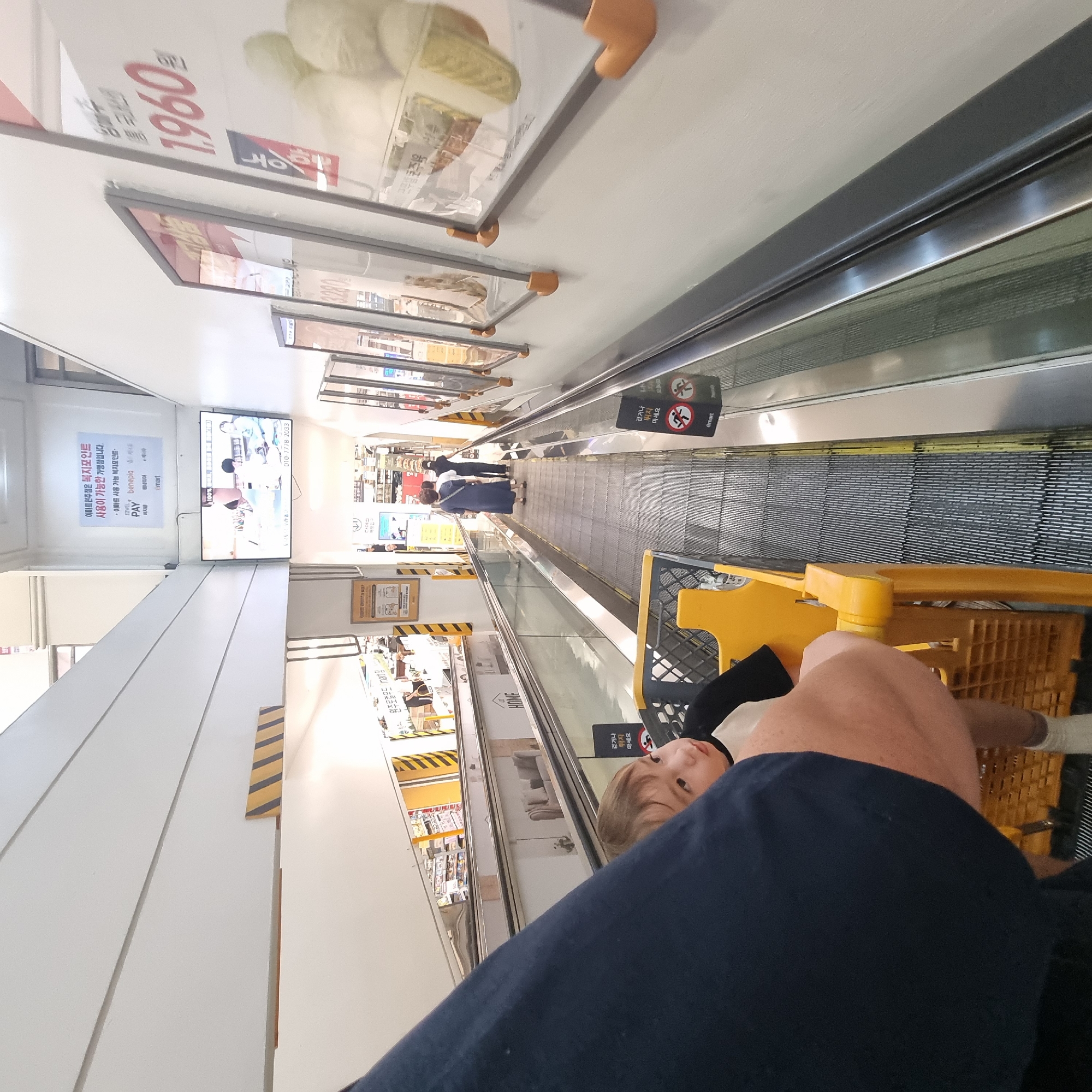 The image shows an indoor escalator in a retail environment, likely a department store or supermarket, given the presence of promotional displays and product information on the walls. The escalator is moving, and a young child is seated in a yellow shopping cart next to the camera, looking to the right. The child is wearing a black and white shirt and white socks. In the background, several adults are visible walking on the escalator. One adult is wearing a blue dress, and another is wearing black clothing. Promotional signs are displayed on the walls alongside the escalator, with one sign showing a price of "1,960". Another sign appears to be an advertisement for "e•mart". The escalator is enclosed by glass railings. The overall environment suggests a commercial space with a focus on shopping. The mention of "e•mart" suggests the location is a branch of the E-Mart retail chain, which has numerous locations throughout South Korea, including in Wonju-si.