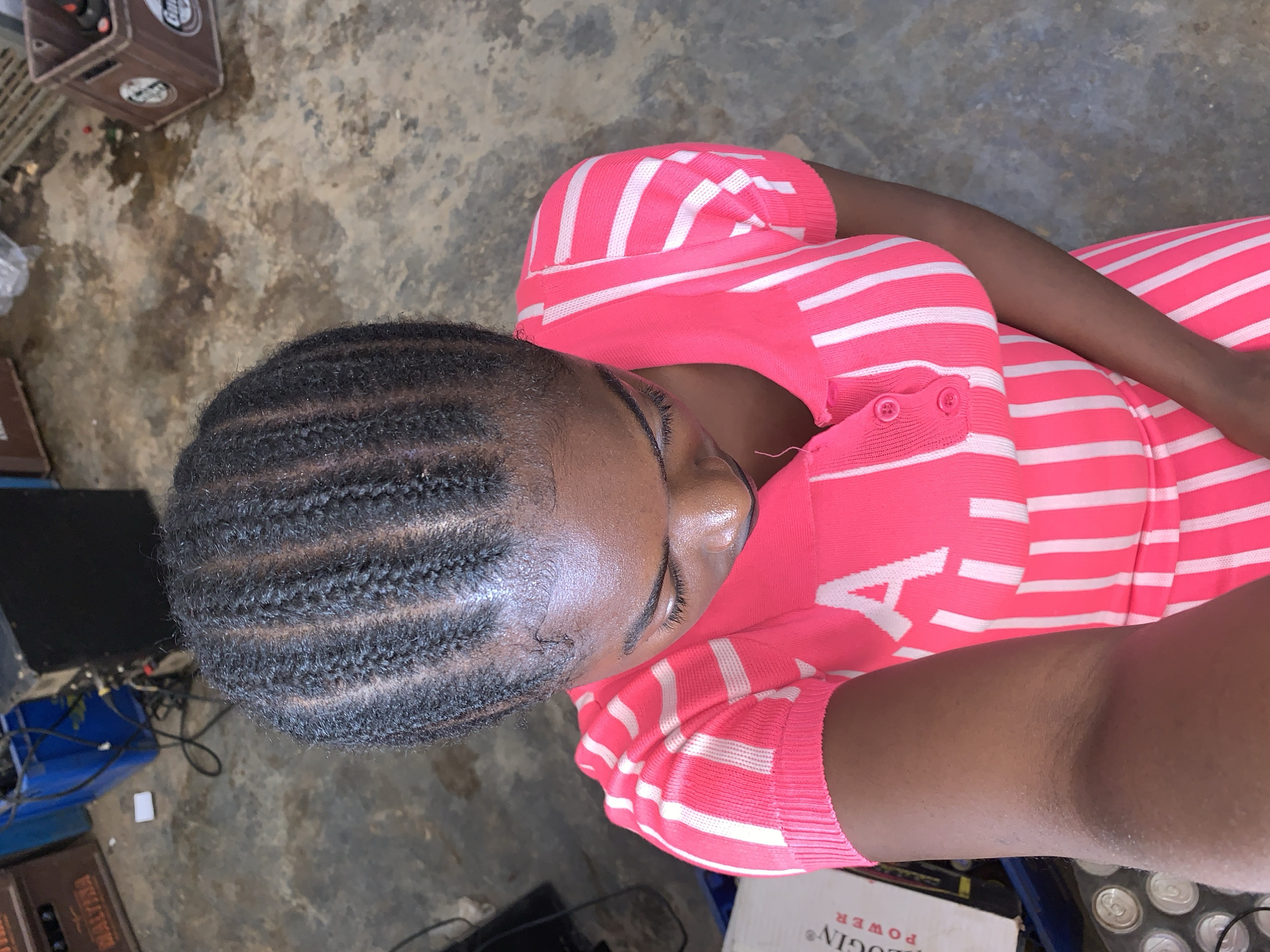 A medium-angle, close-up shot shows a woman lying down, her head and upper body visible. Her hair is styled in close braids, revealing her scalp. She is wearing a pink and white striped sweater with buttons at the shoulder.
The background is a cluttered, gritty area with various objects. To the left, there's a black electronic device with wires, and a blue plastic container. A brown crate is visible in the top left corner and another brown crate in the bottom left. A white box with the word "LOGING" printed on it is in the bottom right. Cans are present next to the box. The ground appears to be concrete.
Given the information, there is no clear indication that this image was taken in the city of Kabong, Nigeria.