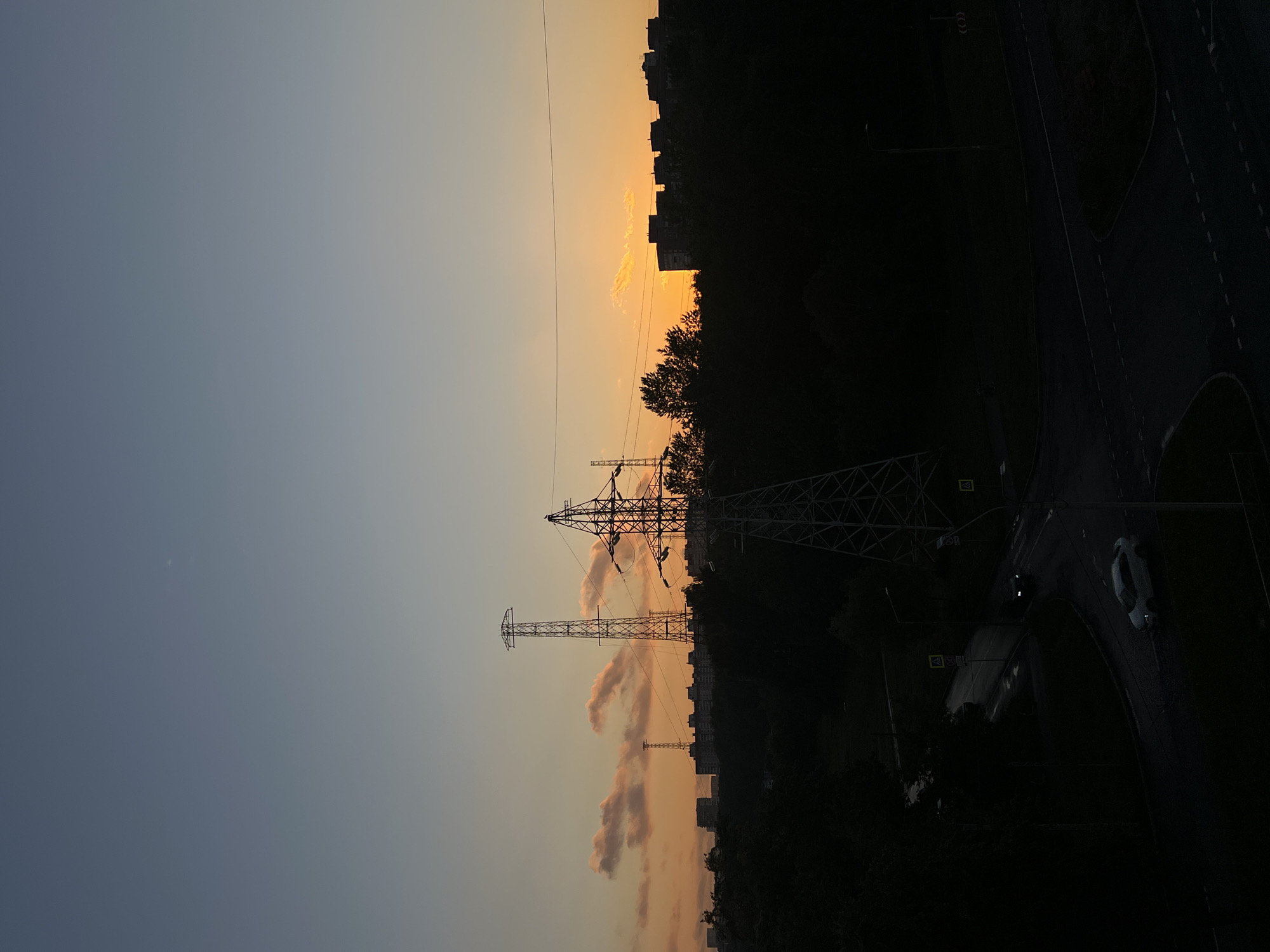This is a vertical, eye-level, medium shot of an urban landscape at sunset. The sky transitions from a deep blue on the left to a warm orange and yellow on the right, with wispy, pink-tinged clouds scattered throughout. Two large electricity pylons dominate the center of the frame, silhouetted against the colorful sky. Behind them, a dark line of trees and buildings runs horizontally across the image. To the far right, a dark road with white lane markings curves away, with a single white car visible on it. The overall mood is one of tranquility and the quiet end of a day, with the dramatic sunset providing a sense of beauty. The setting appears to be a residential area on the outskirts of Saint Petersburg, Russia, evidenced by the apartment blocks and the presence of power lines and infrastructure typical of a large city. The lighting suggests it is late evening, with the sun just below the horizon. There is no visible text in the image.
