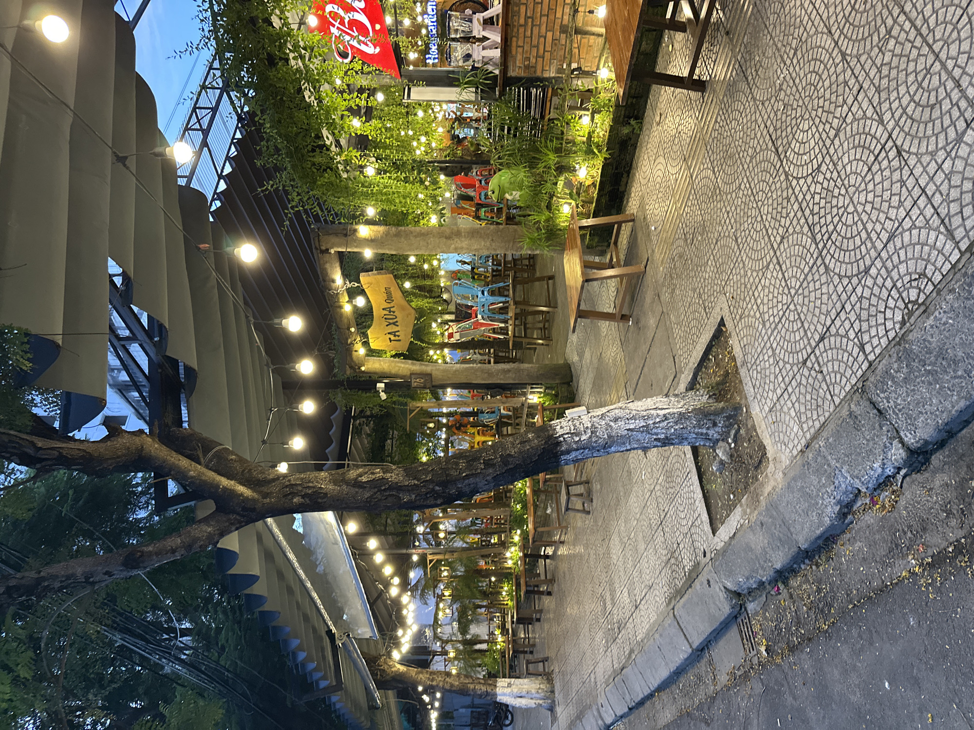 The image depicts an outdoor seating area of a restaurant or cafe in Đà Nẵng, Vietnam. The scene is illuminated by numerous string lights that hang from a canopy structure. Tables and chairs, mostly made of wood and metal, are arranged on a tiled patio. Lush greenery, including hanging plants and potted flora, is integrated into the decor, creating a natural and inviting atmosphere. A prominent sign with Vietnamese lettering, "TÀ XÓA Quán," is visible, indicating the name of the establishment. The overall ambiance suggests a casual dining or gathering spot, likely popular in the evening due to the lighting.