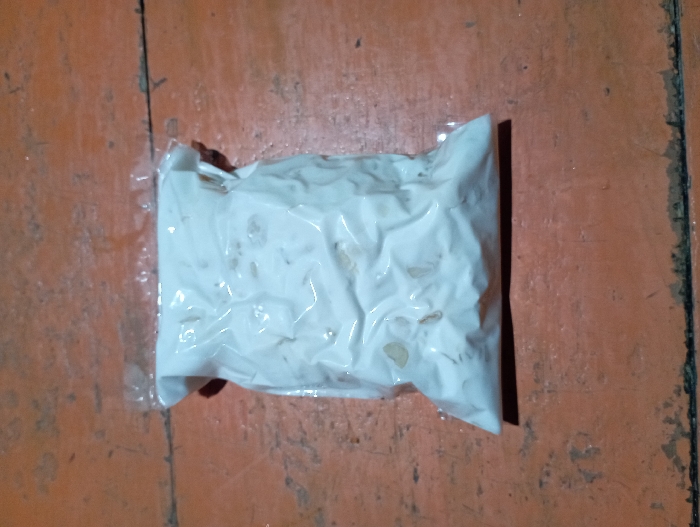 An overhead shot captures a rectangular package of tempeh, a traditional Indonesian fermented soybean product, laid horizontally on a rustic wooden surface. The tempeh itself is a solid, white block with visible pieces of soybeans or grains embedded within the mycelial culture, all encased in a clear, tightly sealed plastic wrapper that conforms to its lumpy shape.

The package rests on an orange-brown wooden surface, likely a floor or tabletop, composed of multiple planks. These planks display distinct grain patterns, several visible cracks, and dark lines marking the joins between them. The wood shows signs of wear and discoloration, suggesting an aged or well-used surface.

No people are present in the image, and no specific activity is taking place; it is a still life of the food item. The lighting appears soft and diffused, indicating an indoor setting. There is no discernible text or branding visible on the packaging or in the background. The scene is set in Pubian, Indonesia.