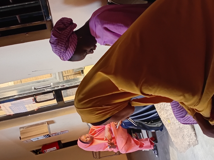 Three women are pictured in a room. One wears a pink hijab, holding an object. Another wears a purple head covering and a light purple outfit. The third woman is wearing a mustard yellow outfit. A window is visible, and a red fire extinguisher is also present.