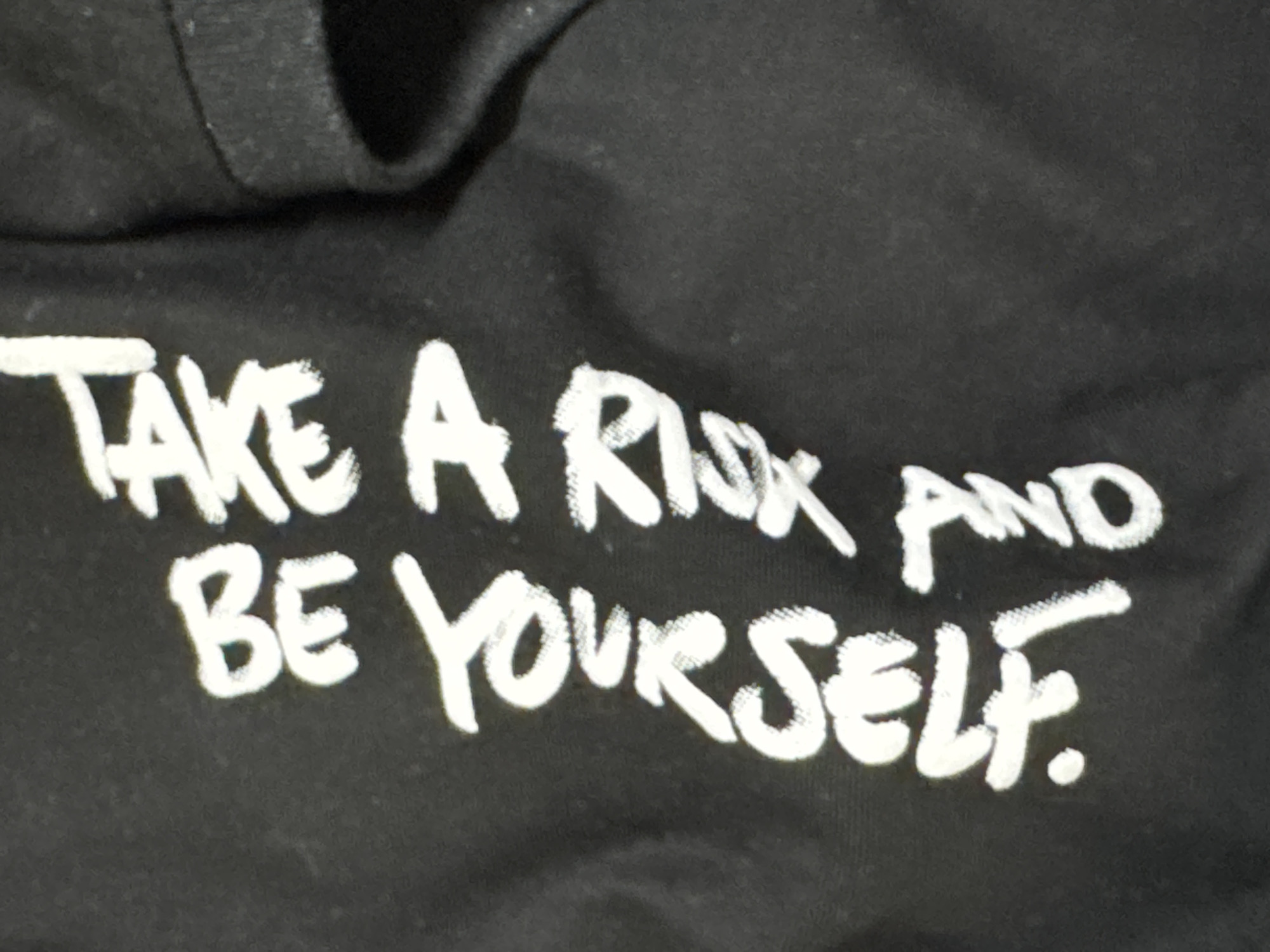 The image shows a close-up of a black garment with white lettering. The text reads "TAKE A RISK AND BE YOURSELF."