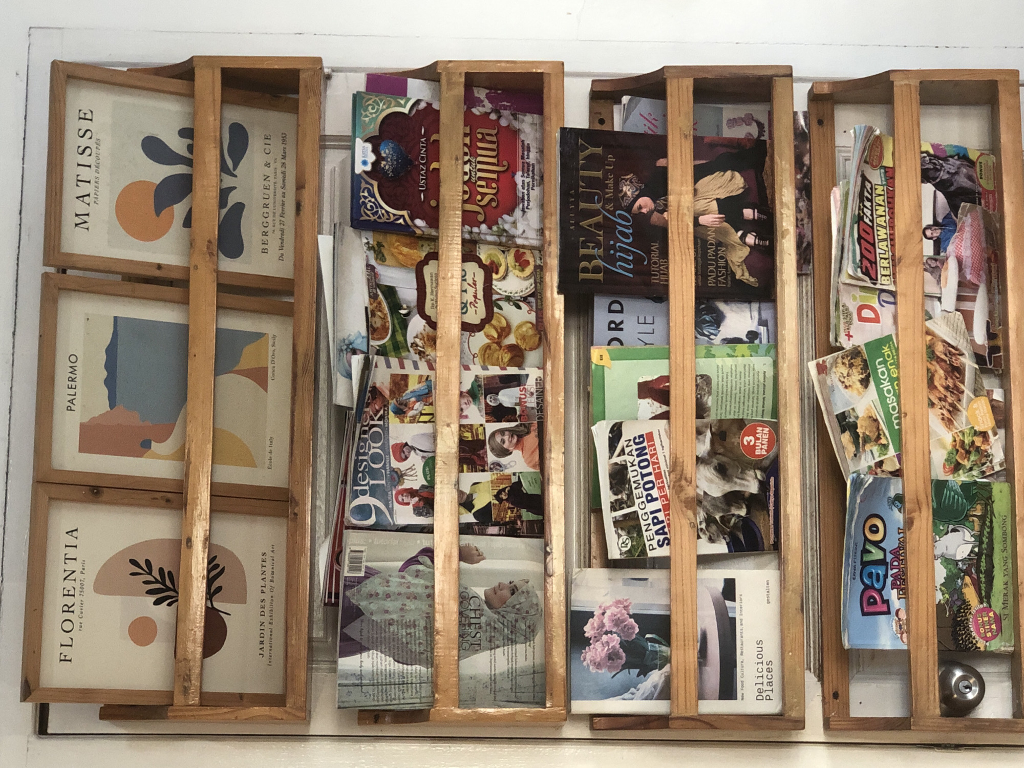 Here is a detailed description of the image:
The image shows a set of four wooden shelving units mounted on a white surface. The shelves are filled with a variety of printed materials, including magazines, books, and framed prints.
The shelf furthest to the left contains three framed prints: "MATISSE PAPIERS DECOUPES", "PALERMO" and "FLORENTIA rue Cuvier 75007, Paris", "JARDIN DES PLANTES International Exhibition Of Botanical Art".
The next shelf contains magazines with images of fashion, food, and models. Some of the magazines appear to be Indonesian publications.
The third shelf contains books and magazines, including a book titled "Beauty hijab TUTORIAL HIJAB & Make Up", and Indonesian magazines about fashion and cooking.
The fourth shelf contains Indonesian cooking magazines. A silver doorknob is visible at the bottom right corner of the shelf.
The subject matter indicates the location is likely a home, store, or office in Indonesia, possibly in Lhokseumawe.