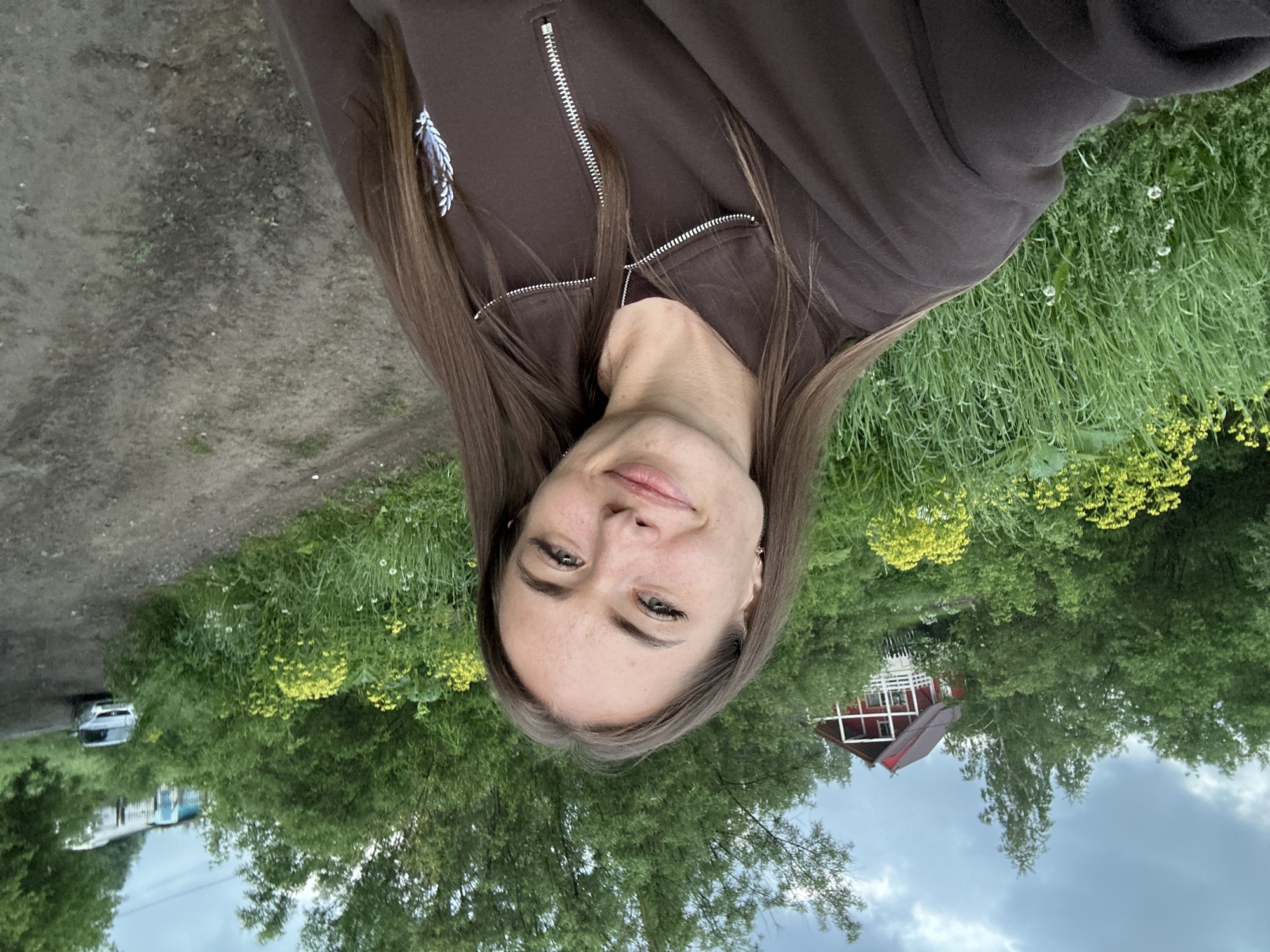 A young woman with long brown hair is taking a selfie outdoors. She is smiling and looking at the camera. She is wearing a brown zip-up sweatshirt. The background is a lush green landscape with trees, yellow flowers, and a dirt road. A red house with white trim is visible in the distance, partially obscured by trees. A silver car is parked on the side of the dirt road. The sky is cloudy. The setting appears to be rural.
