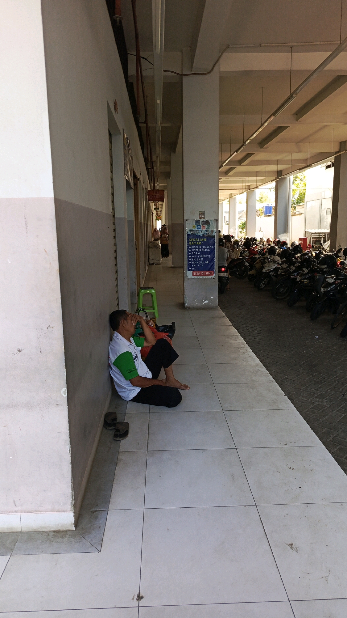 A middle-aged man with dark hair is sitting on the tiled ground, leaning against a white wall. He is wearing a white polo shirt with green accents on the sleeves and collar, and dark pants. His right hand is covering his forehead as if he is stressed or tired. A black bag is placed on the ground behind him, and a pair of brown sandals are next to his feet. A green plastic stool is positioned a few feet away from the man. The scene appears to be an outdoor corridor or walkway, with a row of shops or offices on the left and a parking area filled with motorcycles on the right. A sign on a pillar in the background lists various payment options, indicating a commercial or public area. The overall environment suggests a hot and sunny day. This location is identified as Nologaten, Indonesia.