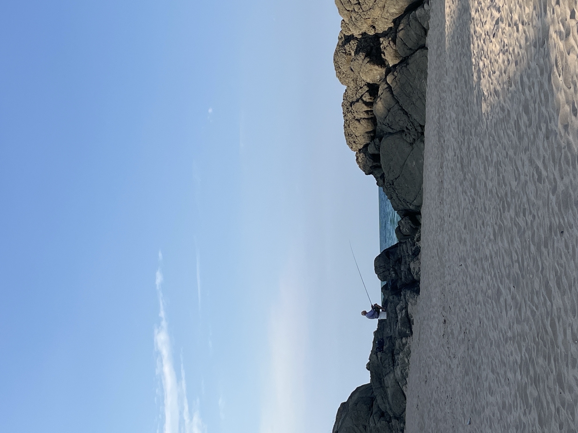 The image depicts a serene beach scene in Ahtopol, Bulgaria. In the foreground, a sandy beach stretches across the frame, showing gentle undulations from the wind or tides. Dominating the midground are large, grey, rugged rocks, forming a natural barrier between the beach and the sea. The rocks are textured and weathered, with visible crevices and shadows.
Between the rocky outcrops, a sliver of the calm, turquoise sea is visible, meeting a hazy horizon. A single individual, a man, is seated on the rocks on the left side of the image. He is wearing a light-colored shirt and appears to be fishing, holding a long fishing rod that extends out over the water. Next to him sits a white bucket.
The sky above is a clear, pale blue, with a few wispy white clouds scattered towards the left. The lighting suggests it is either early morning or late afternoon, with a soft, diffused sunlight casting subtle shadows. The overall atmosphere is one of peaceful solitude and a connection with nature.