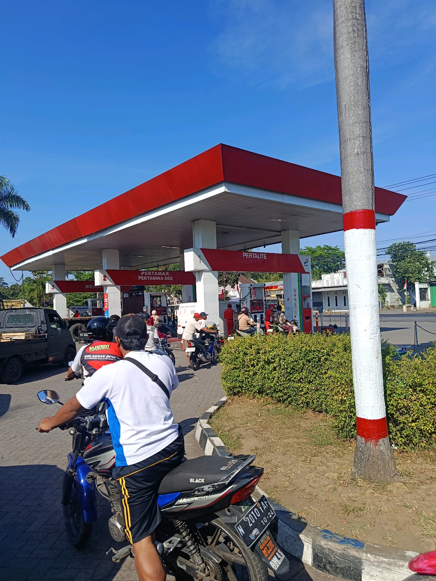 The image captures a sunny day at a Pertamina gas station in Tempeh Lor, Indonesia. In the foreground, a person is seated on a motorcycle, viewed from behind. The motorcycle has the license plate "N 2010 YAF". The gas station itself is visible, with its distinctive red roof and white support pillars. Fuel pumps are present, and several other motorcycles and a small truck are also in the vicinity, indicating activity at the station. People are seen at the pumps, and some are standing around. A prominent concrete pole, painted with red and white bands, stands to the right of the frame. Lush green bushes border the paved area of the gas station. The sky is clear and blue.