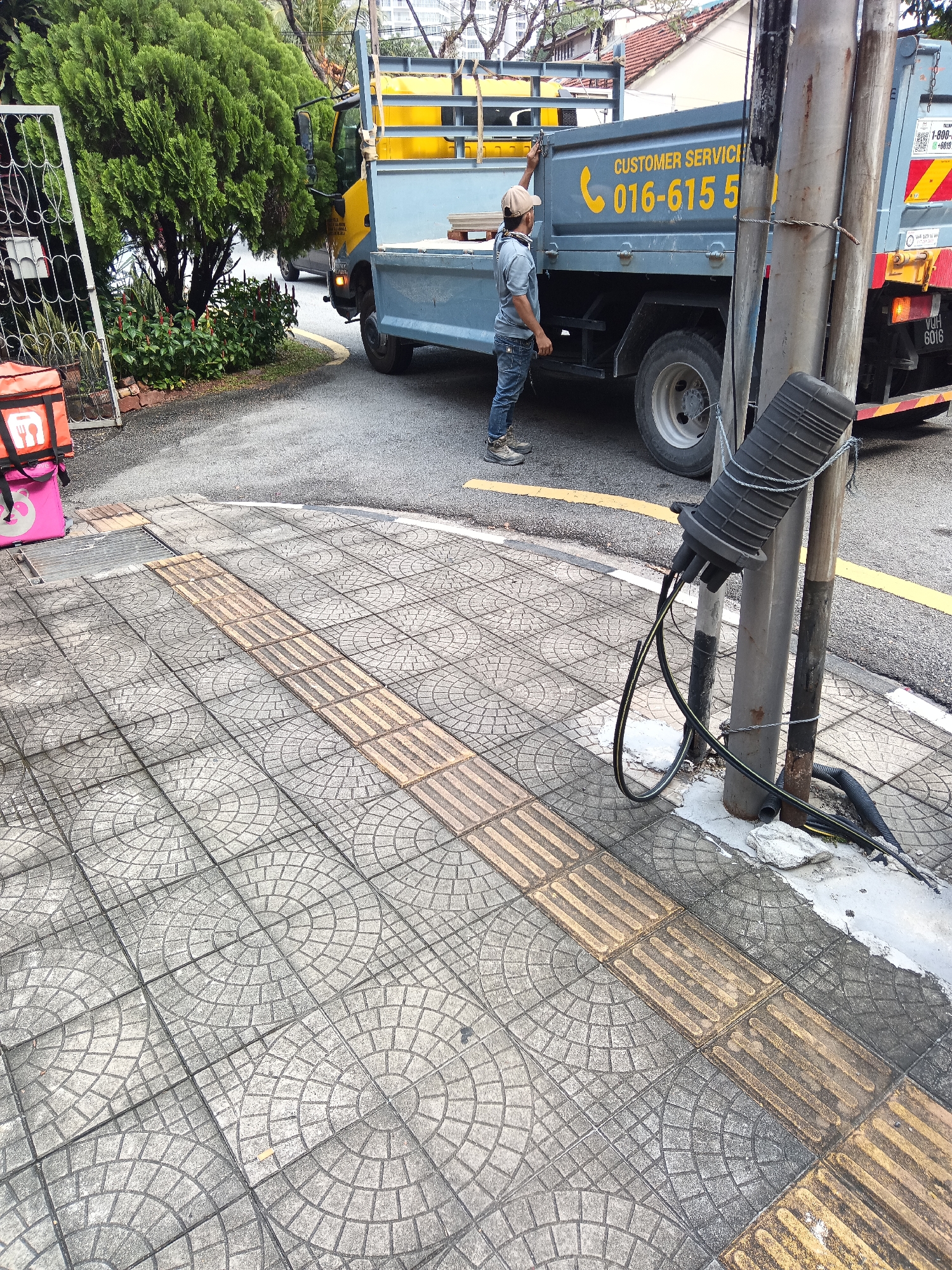 The image shows a street scene in Kuala Lumpur, Malaysia. In the foreground, a tactile paving sidewalk is visible, marked with a tactile guidance path. To the right, a utility pole with attached cables and conduit stands near the sidewalk. Behind the sidewalk, a light blue truck with yellow accents is parked on the street. A person wearing a cap and blue jeans is standing next to the truck, appearing to be interacting with it. An orange insulated food delivery bag with a white logo is placed on the ground to the left of the sidewalk, near a white metal gate and lush green trees. The street has a yellow line marking.