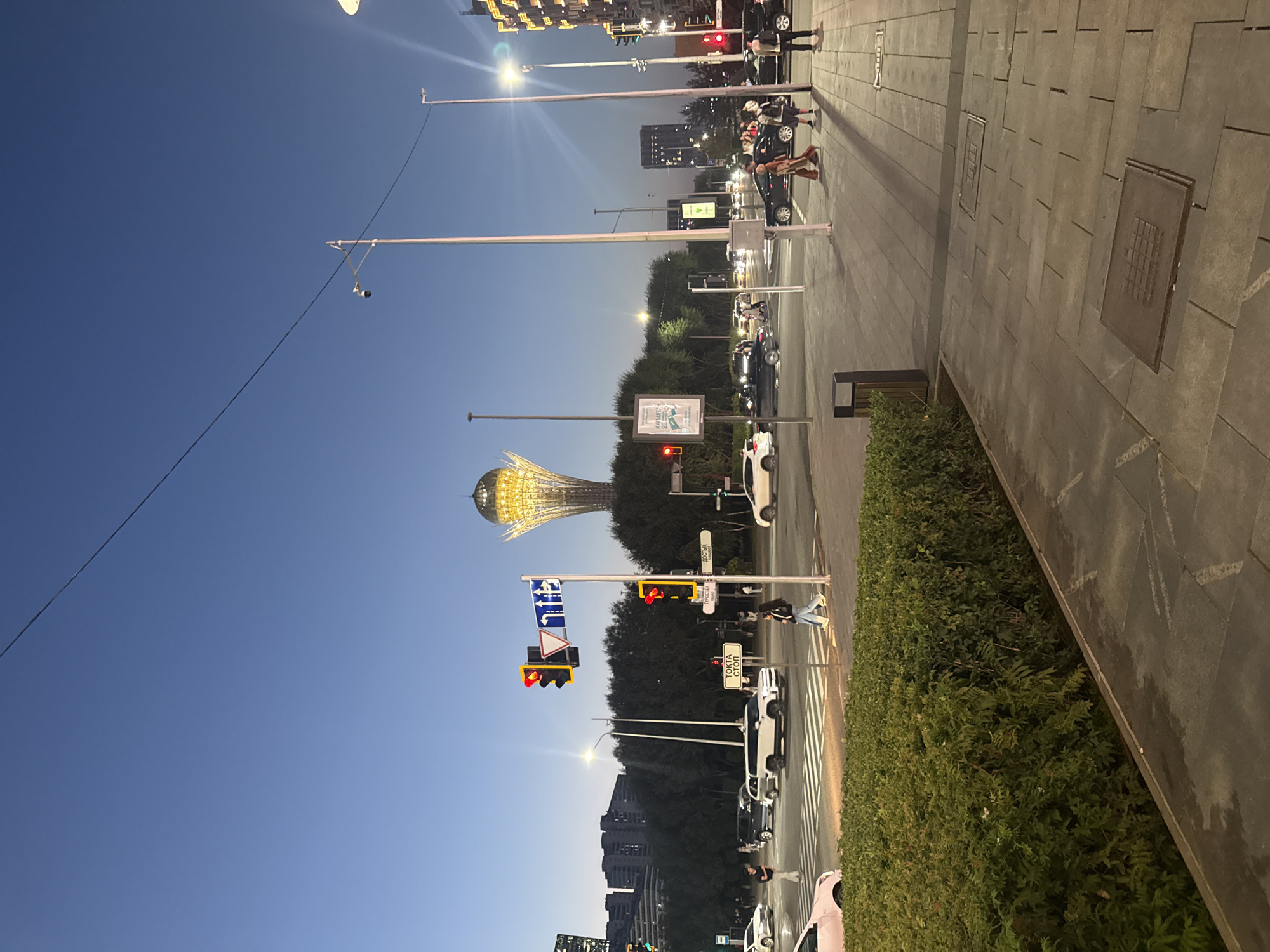 An urban street scene at dusk in Astana, Kazakhstan, is depicted. The iconic Bayterek Tower, illuminated with golden lights, stands prominently in the background against a clear blue sky transitioning to darkness. The foreground features a pedestrian crossing and sidewalk, with a lush green hedge lining the left side. Several cars are visible on the road, some stopped at a traffic light which displays a red signal. Pedestrians are crossing the street and walking on the sidewalk. Streetlights and traffic lights are illuminated. Buildings, including a modern high-rise, are visible in the distance on both sides of the street. The overall atmosphere suggests a city street during evening hours.