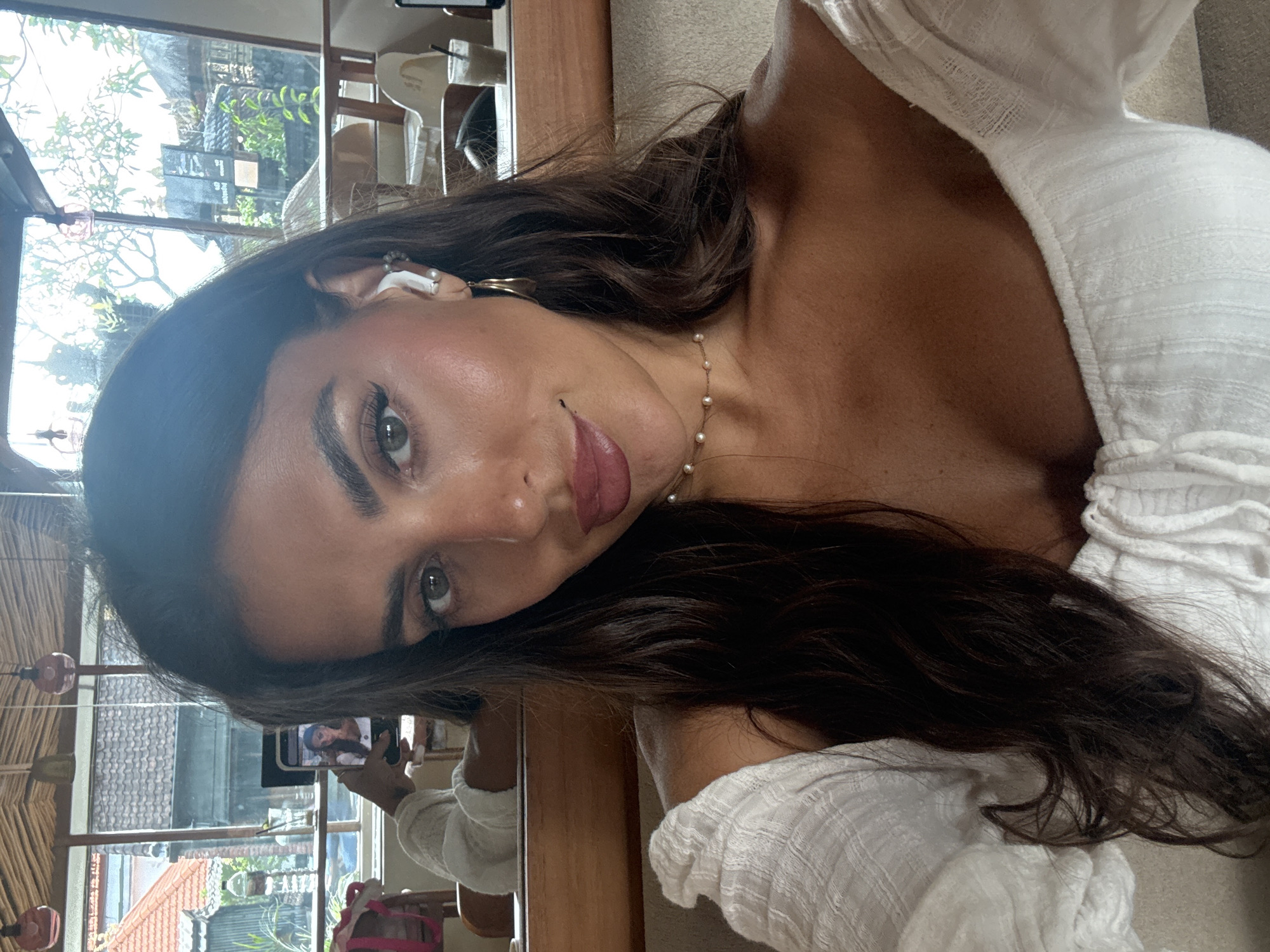 A medium, close-up, portrait-style photograph depicts a woman indoors in North Kuta, Indonesia. Her face is angled toward the viewer. The woman has long, dark brown hair, olive skin, and green eyes. She is wearing a pearl necklace and a white top. An earbud is visible in her left ear. She is holding up a smartphone to take the photo, and the phone's screen shows a smaller image of her taking a photo of herself. The background shows a window with foliage outside, and parts of the interior of what appears to be a cafe or restaurant. There is a table and chairs visible.