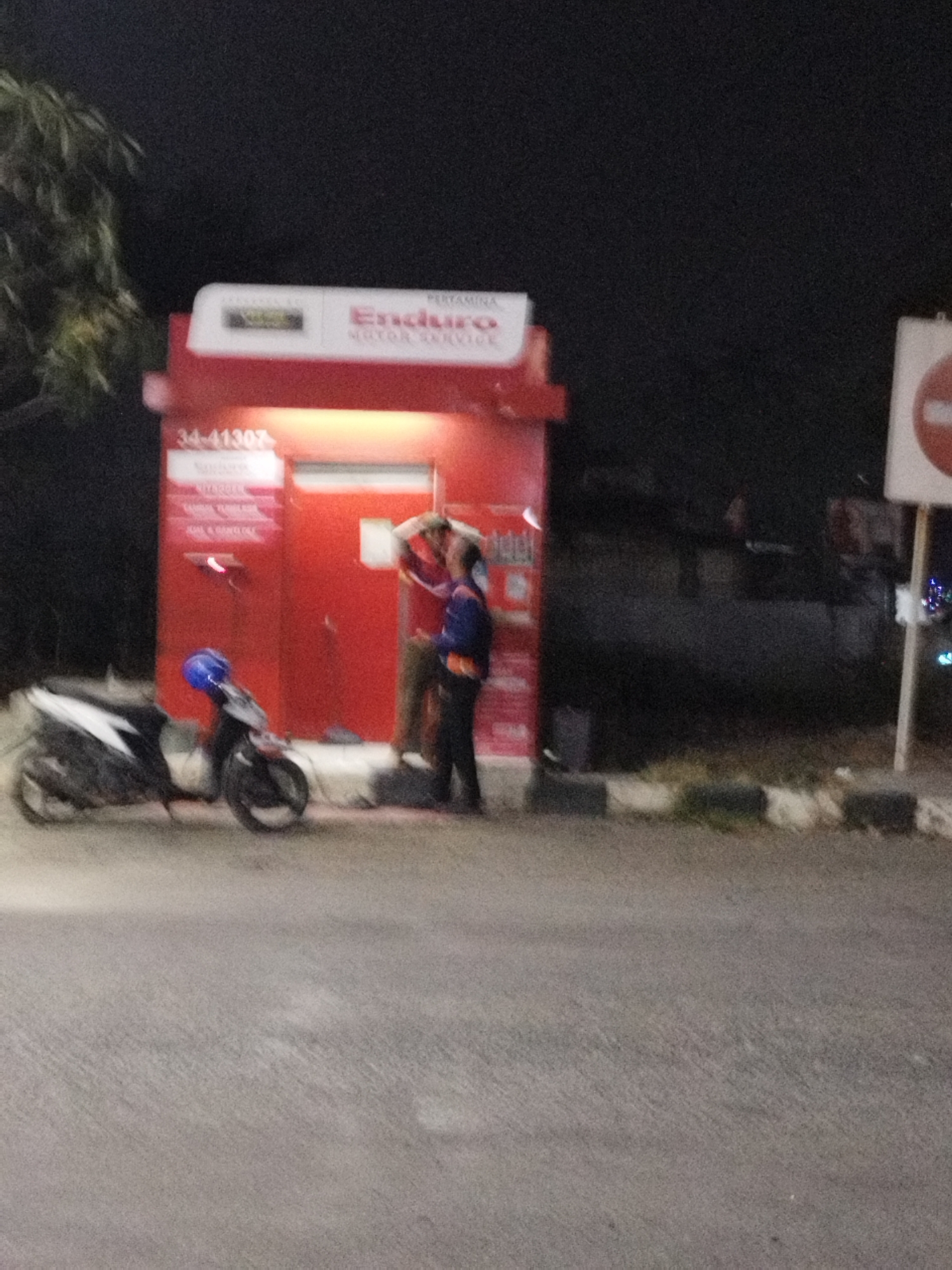 The image depicts a night scene in Karawang, Indonesia, near a Pertamina Enduro gas station. Two individuals are standing in front of a red structure, which appears to be a service kiosk. One person, wearing a blue jacket and a dark shirt, is leaning against the structure. The other person, wearing a red and white shirt, is standing with their arms raised, seemingly engaged in conversation or an activity with the first individual. A motorcycle is parked to the left of the kiosk, its front wheel turned towards the left. The kiosk itself has text and numbers visible on its surface, including "34-41307" and "Enduro MOTOR SERVICE." The area is dimly lit, with artificial lighting illuminating the kiosk and surrounding pavement. The background is dark, suggesting it is nighttime.