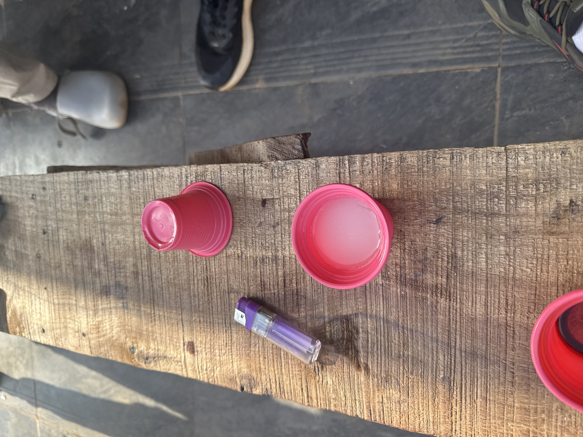 The image shows a weathered wooden surface, possibly a bench or table, with several items on it. There are three red plastic cups, one overturned, one filled with a translucent liquid, and one partially visible at the edge of the frame. A clear plastic lighter with a purple top lies between two of the cups. Parts of footwear belonging to unseen people are visible at the top of the image. The image was taken in Kurunduma, Nigeria.