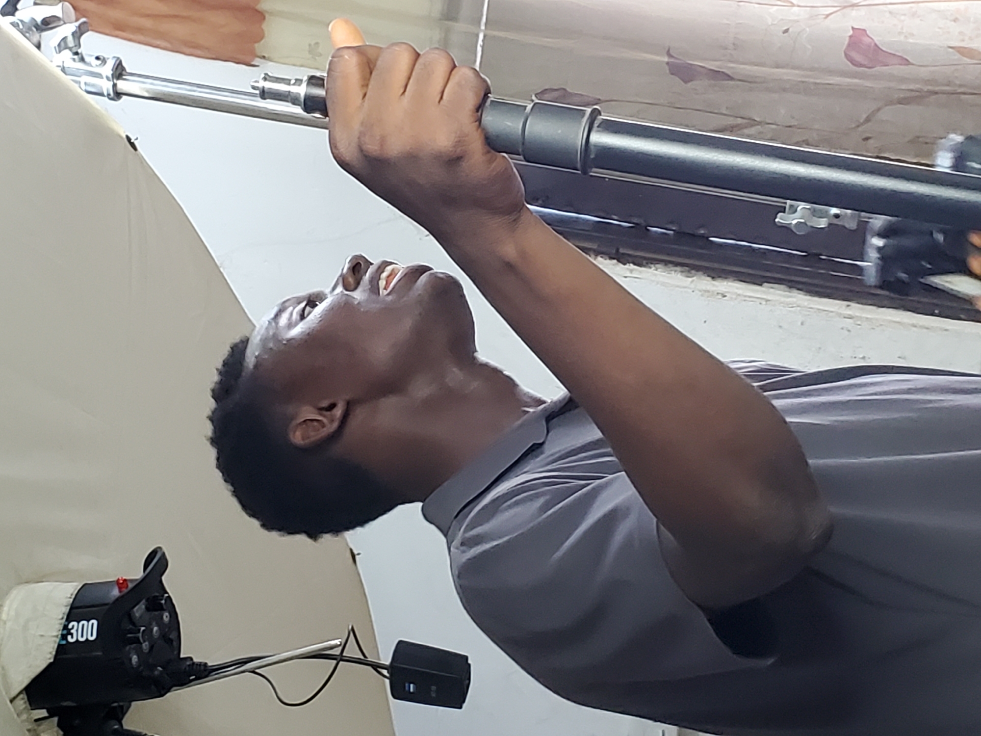A person is holding onto a metal rod above them, likely adjusting studio lighting equipment. The person has dark skin, short black hair, and is wearing a gray shirt. The lighting equipment includes a large white diffuser panel and a black device labeled "300," which is likely a studio flash or strobe unit. The background includes a curtain or window covering. The location is presumed to be Mararaba, Nigeria.