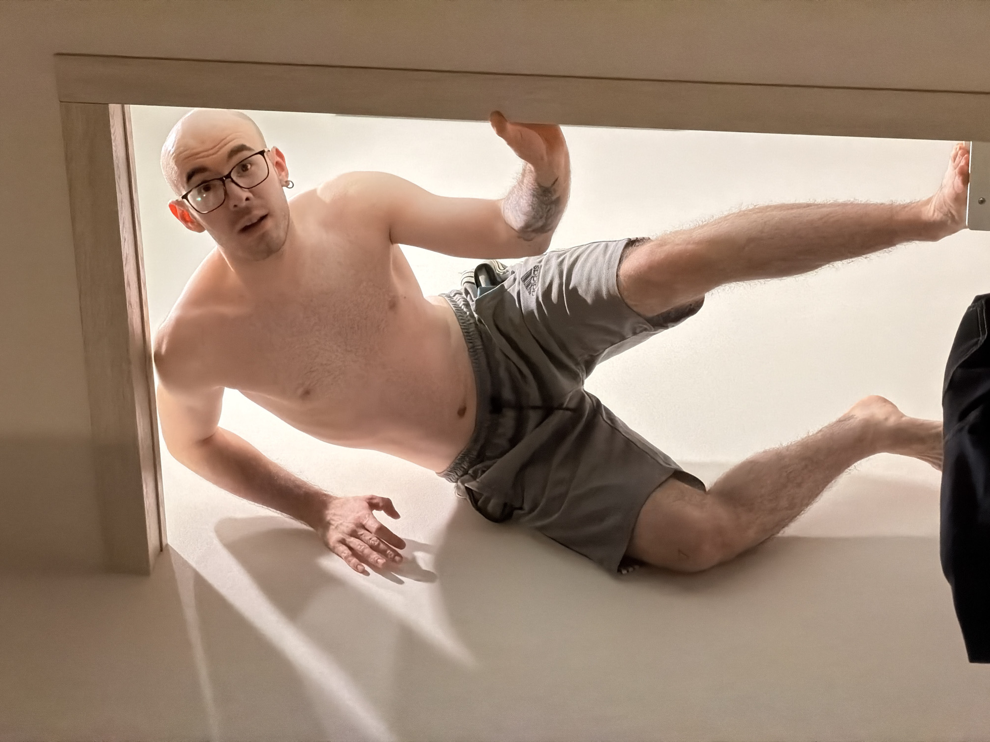 A light-skinned, bald man with glasses is captured in a photograph. He is lying on a white surface, appearing to be a floor, within a doorframe. The man is wearing gray shorts and is shirtless. His left leg is extended straight out, with his foot touching the upper right corner of the doorframe. His right leg is bent at the knee, with his foot on the floor. His right arm is raised with his palm facing outward, while his left arm is extended towards the floor. The location of Kazan, Russia, is not discernible from this image.