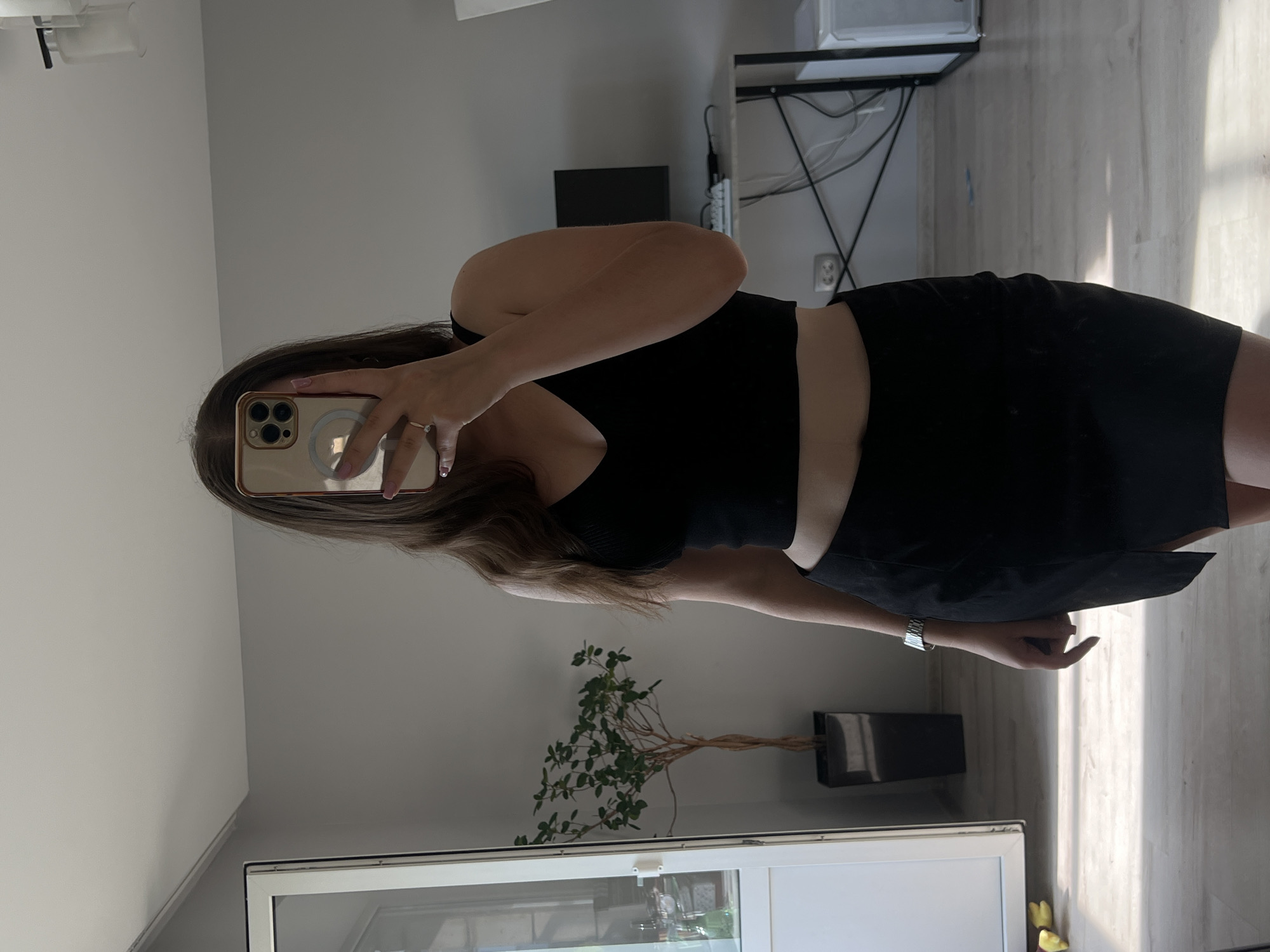 A young woman, with long brown hair, stands in a room and takes a mirror selfie. She is wearing a black crop top and a black mini skirt. The background of the image shows a grey wall, a large mirror, a potted plant, a desk with a computer monitor, and a refrigerator. There is also a door with glass panels visible on the left side of the image. The room appears to be in an apartment in Ternopil, Ukraine.