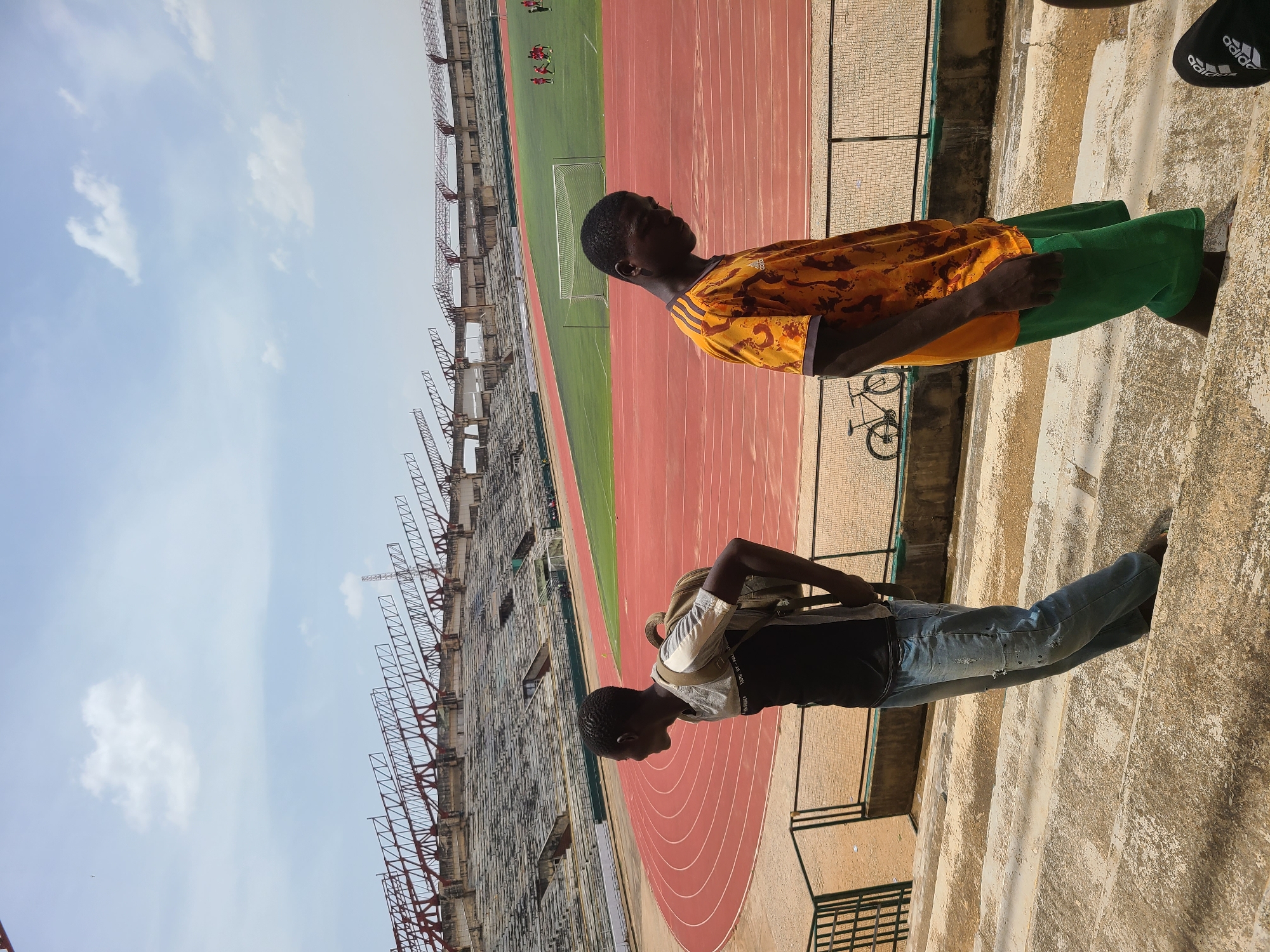 The image captures an elevated view of a sports stadium under a bright, partly cloudy sky, consistent with the "New Stadium, Nigeria" context. Two young men are positioned on concrete steps or a walkway, observing the activities below.
In the foreground, a young man wears a light grey short-sleeved top over a black t-shirt, blue jeans, and carries a light brown backpack. White text reading "EVERY DAY" is visible on his black t-shirt. He is looking towards the left side of the frame, down into the stadium. To his right and slightly further back, another young man stands, dressed in a vibrant yellow and orange patterned short-sleeved shirt, which features black Adidas stripes on the sleeves, and green shorts. He is also looking into the stadium.
Below the observers, a red running track encircles a lush green football pitch. A white goalpost is visible on the pitch, and several small figures, likely football players, can be seen in the distance, suggesting a match or training session is in progress. The stadium's stands or seating areas rise in the background, characterized by concrete structures and exposed metal frameworks.
Notable objects include a black bicycle partially visible behind the man in the yellow shirt, and a pair of black flip-flops with "Adidas" branding on them, seen near the feet of the man in the yellow shirt. The bright sunlight indicates it is daytime, casting distinct shadows. The overall scene depicts a casual observation of a sporting event or practice at a stadium.