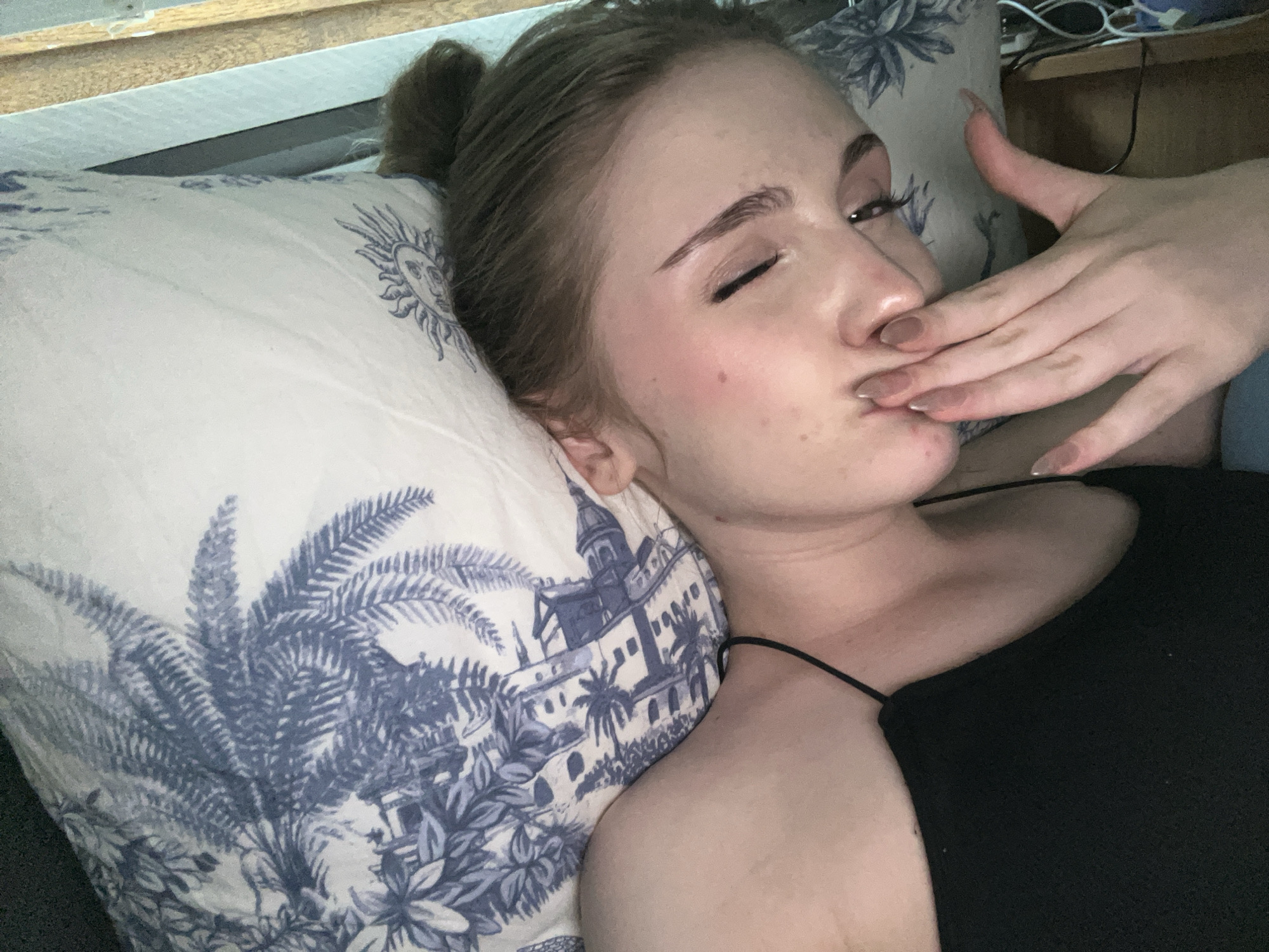 The image shows a young woman lying on a pillow with a blue and white pattern. She has her hair pulled up in a bun and is wearing a black top. Her left eye is closed, and her right hand is covering her mouth. The pillow has a toile-like print featuring trees and buildings. The background appears to be a room with wooden furniture and some electronic equipment. There is no information available about the location being Ashkadar, Russia.