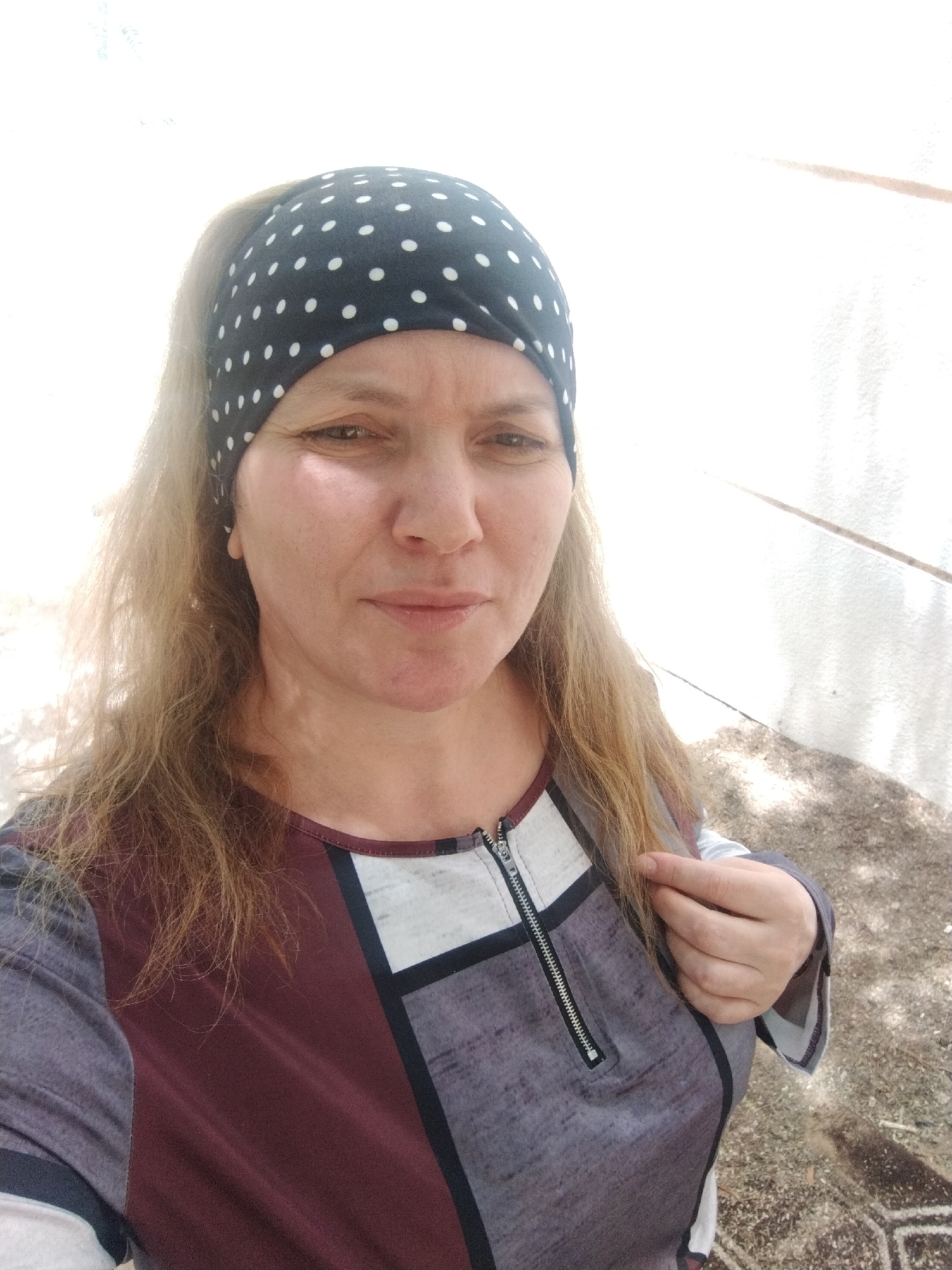 A middle-aged woman with light brown, shoulder-length hair is wearing a black and white polka dot headband and a patterned long-sleeved shirt. The shirt has a maroon section, a grey section, and a white square around the neck with a zipper detail. She is standing outdoors in bright sunlight. The background shows a white wall with an area of dirt and sparse vegetation. The image was captured in the city of Al Mas'oudi, United Arab Emirates.