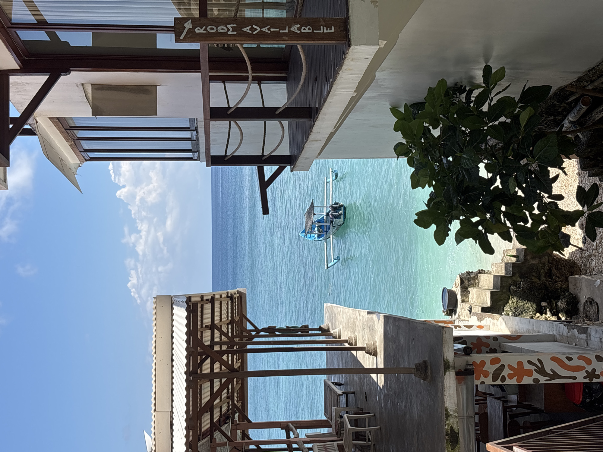 The image captures an ocean view in Uluwatu, Indonesia, likely taken from an elevated position, possibly a hotel or guesthouse. The perspective is somewhat tilted. The foreground features architectural elements of the building: wooden beams, a white awning, and sections of walls. A partially obscured sign reads "ROOM AVAILABLE" (the spelling is slightly altered). A lush green tree is on the right side.
In the middle ground, a small boat with a blue and white canopy is moored in the clear turquoise water. Beyond the boat, the ocean extends to the horizon.
The sky is blue, with a few white clouds visible to the left of the frame. The photograph appears to be taken during the day, with natural sunlight illuminating the scene. There are no people visible in the image.