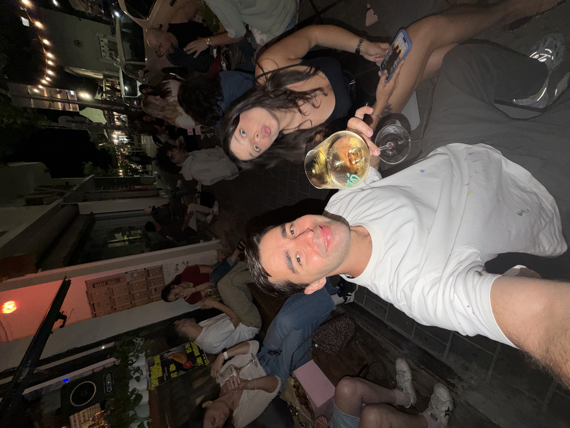 A group of people gather at night, possibly in an outdoor setting, in Tel-Aviv, Israel. Some are seated on the floor, while others stand. A man in a white shirt holds a glass of wine, posing for a photo with a woman. The scene is lit by string lights overhead, with a building in the background. Other individuals are scattered around, some looking at their phones. The environment appears casual and social.