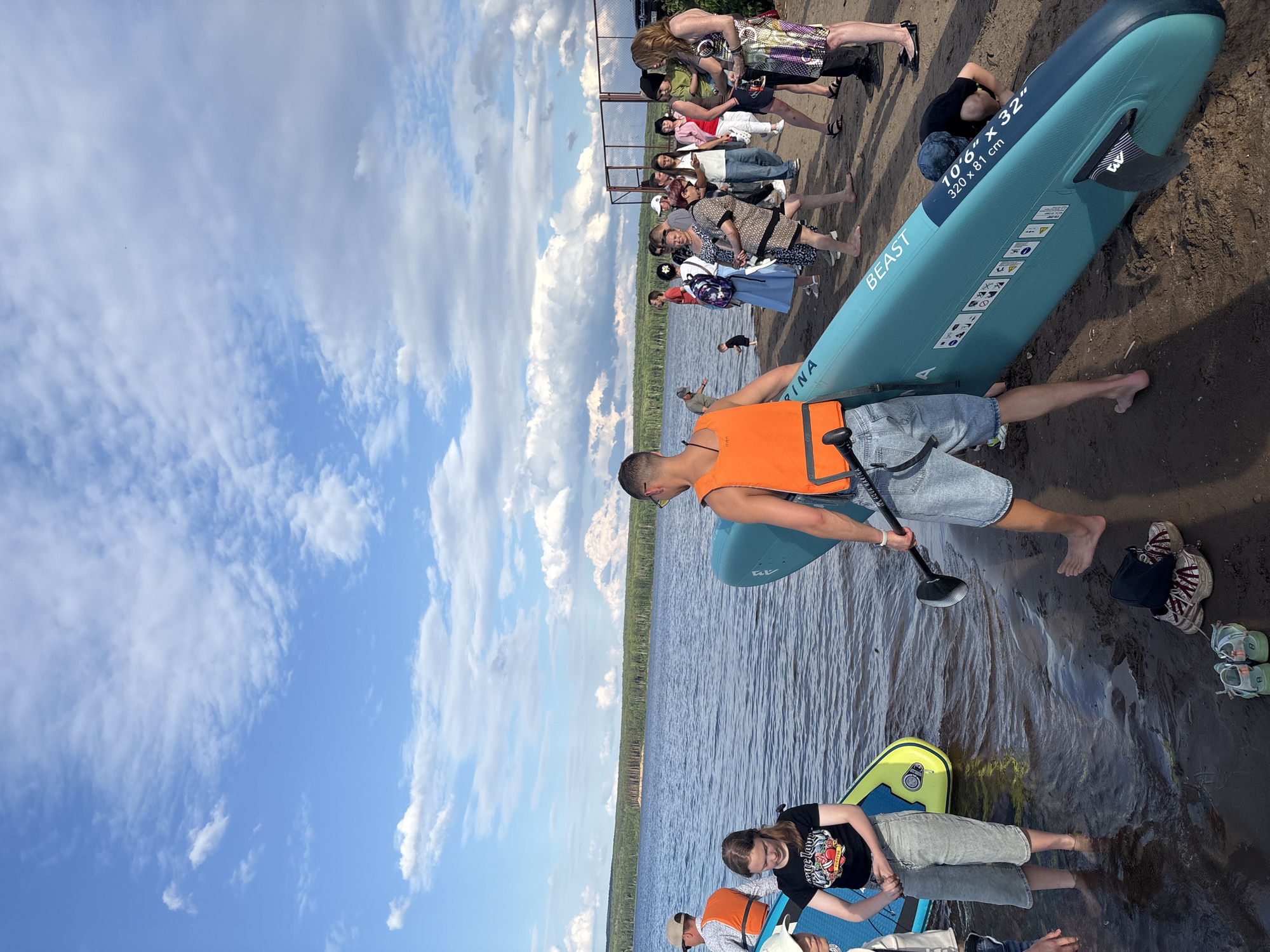 The image depicts a group of people gathered on the sandy shore of the Kama River in Chaikovsky, Russia, preparing for or having just concluded stand-up paddleboarding activities. The sky is partly cloudy with a vibrant blue expanse. In the foreground, a man wearing an orange life vest and denim shorts holds a paddle, standing next to a teal stand-up paddleboard labeled "BEAST 10'6" x 32"". To his left, a young girl in a black t-shirt and light-colored capris stands in the shallow water, with another paddleboard partially visible. Further back, a diverse group of individuals, some wearing life vests and others in casual attire, are congregated on the beach. The background features the calm waters of the Kama River, bordered by a dense line of trees, and a metal fence structure. The scene appears to be a recreational outing on a warm day.