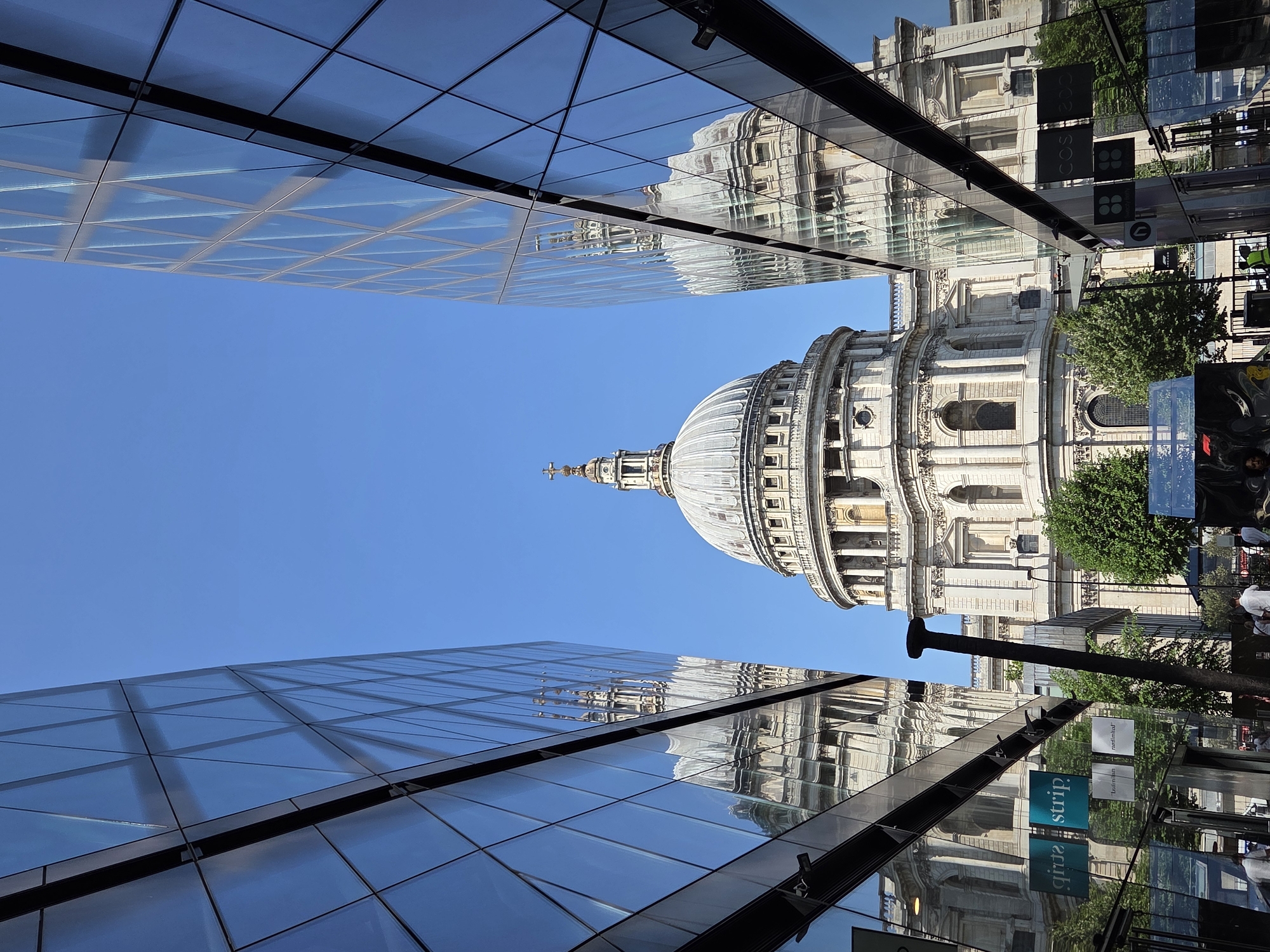 The image is a low-angle view of St. Paul's Cathedral in London, framed by modern glass-clad buildings. The cathedral's dome dominates the center of the frame against a clear blue sky. The modern buildings flank the cathedral, their glass facades reflecting both the cathedral and the surrounding environment. Lower right side of the image, there is a street, with people, street signs and businesses, which appear upside down in the reflections.