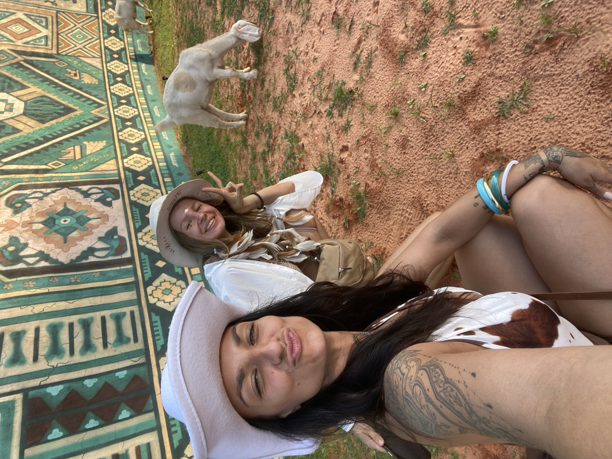 Two women are photographed in Mui Ne, Vietnam, in front of a colorful, tapestry-like mural depicting geometric patterns and stylized figures. The woman in the foreground, with dark hair and wearing a white cowboy hat and a cow-print bikini top, is looking directly at the camera and making a duck-face. She has extensive tattoos on her left arm and shoulder. The woman in the background, also wearing a white cowboy hat, has blonde hair and is smiling at the camera, holding up two fingers in a peace sign. She is wearing a white top with feather-like embellishments around the neckline and a tan crossbody bag. Behind them, two goats are visible on a patch of grass. The ground in the foreground appears to be reddish sand with sparse green vegetation. The overall setting suggests an outdoor, possibly festive or tourist-oriented location in Mui Ne.