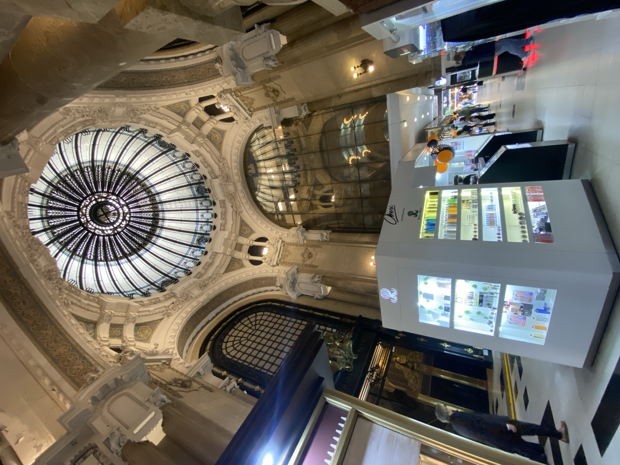 The image shows an interior view of Galerías Pacífico in Buenos Aires, Argentina. A large, ornate dome with a radial pattern of black and white is visible at the top of the image. Below the dome are arched windows. There are shopping stalls on the ground level. A display case stocked with products, lit from within, dominates the right foreground. People are seen walking along the corridor in the background. The architecture is classical, with ornate detailing.