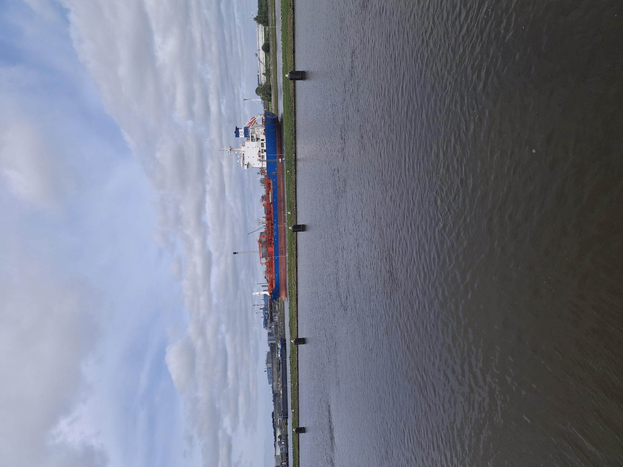 A long, blue and red ship is docked in a calm body of water. The ship has a white superstructure and a tall red and blue smokestack. In the background, there are industrial buildings and a wind turbine. The sky is cloudy and overcast. The water is dark and reflects the sky. There are four black bollards in the water in the foreground.