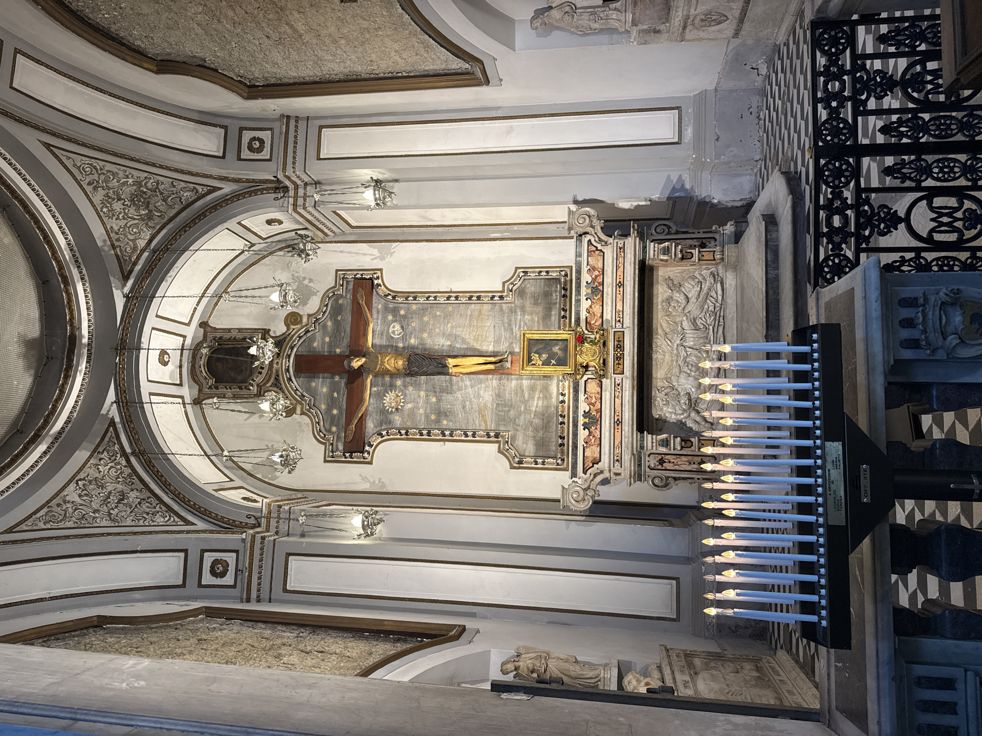 This is a full-shot interior photograph of a chapel or small church, focusing on an altar area. The altar features a large crucifix with Jesus Christ prominently displayed. Above the crucifix is a painting or mosaic. Below the crucifix, there's a smaller framed image and floral decorations. The altar itself is adorned with bas-relief sculptures depicting figures.
In the foreground, a black stand holds numerous lit votive candles. The chapel's architecture includes arched ceilings with decorative moldings and painted details, as well as columns and recessed niches containing statues of figures that appear to be angels or saints. The floor is a checkered pattern of black and white tiles. The overall impression is one of solemnity and religious devotion.
The image is located within the Church of Santi Severino e Sossio in Naples, Italy, specifically in the chapel of the Assumption of Mary.
