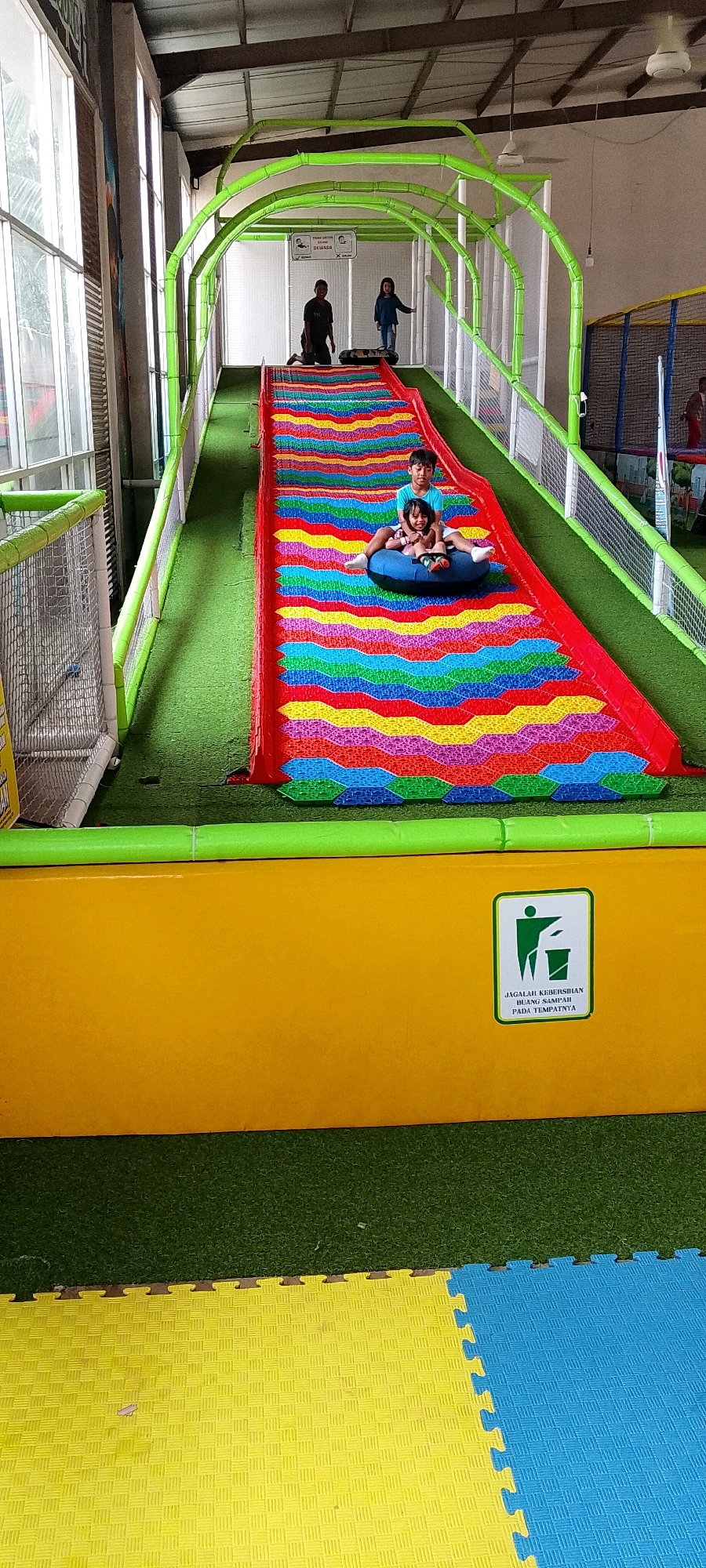 The image captures an indoor playground at Kidzooona in Sidoarjo, Indonesia. The central focus is a brightly colored, rainbow-patterned slide with two children, a boy and a girl, riding down it on a blue inner tube. The slide is enclosed by a green tubular structure. To the left, there's a large window and part of another play area with a netted enclosure. In the background, two adults and a child are visible near the top of the slide, with a sign that reads "AWAS UNTUK DEWASA" (Caution for Adults). The floor is covered with green artificial turf leading to the slide and yellow and blue puzzle mats at the bottom. A yellow barrier with a sign encouraging proper waste disposal is prominently positioned in the foreground.