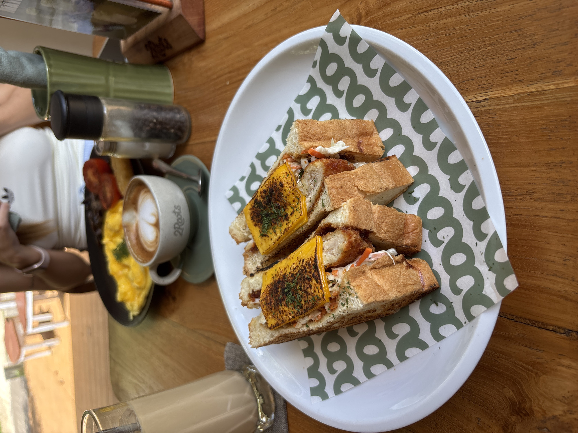 A top-down, slightly angled shot depicts a breakfast or brunch scene on a wooden table. The primary focus is a white plate lined with patterned green and white paper, holding a cut sandwich. The sandwich features toasted bread, a filling of shredded vegetables, a protein (possibly fried chicken or fish), and a slice of melted cheese, possibly cheddar, with a burnt or charred appearance. To the left of the plate, there is a cup of coffee with latte art, next to a small plate of scrambled eggs. A salt and pepper shaker set is also present. Part of a person's arm and clothing is visible on the left side of the frame. A glass containing a beverage (likely a latte or iced coffee) sits in the lower left corner. The location is Pecatu, Indonesia.
